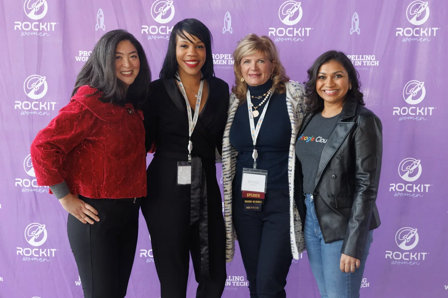 Four women standing together at a conference, smiling for the camera, with a purple backdrop featuring 'Rockit Women' logos and rocket illustrations.