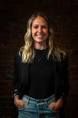 Image of a woman with long blonde hair, with her hands in her pockets, smiling at the camera
