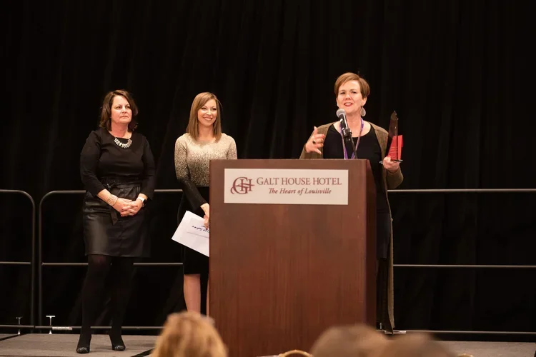 Three women standing on stage at a podium during an award ceremony at the Galt House Hotel, with the woman on the right speaking into a microphone.