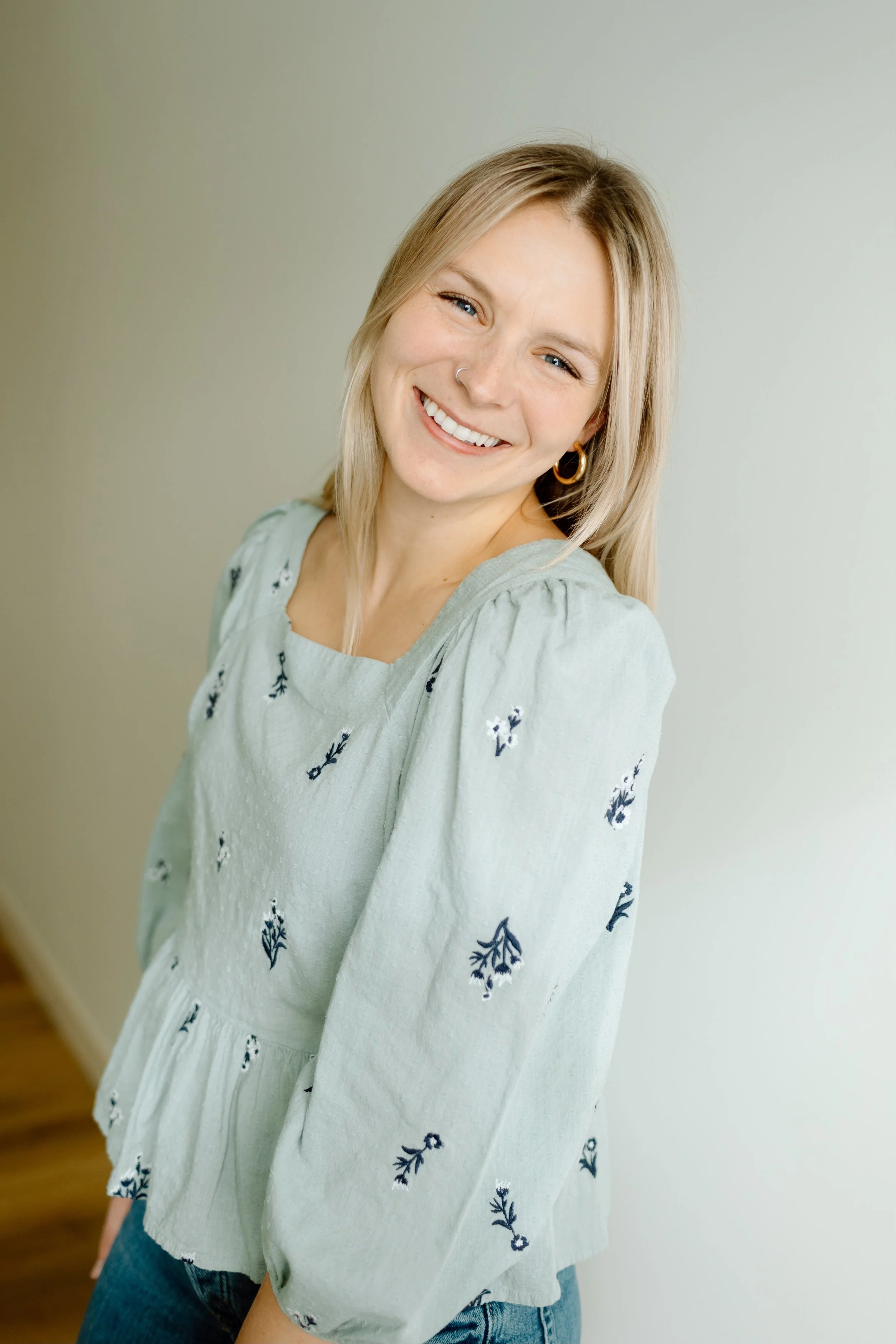 A young woman with blonde hair, wearing a light green blouse with navy floral embroidery, smiling and looking at the camera.