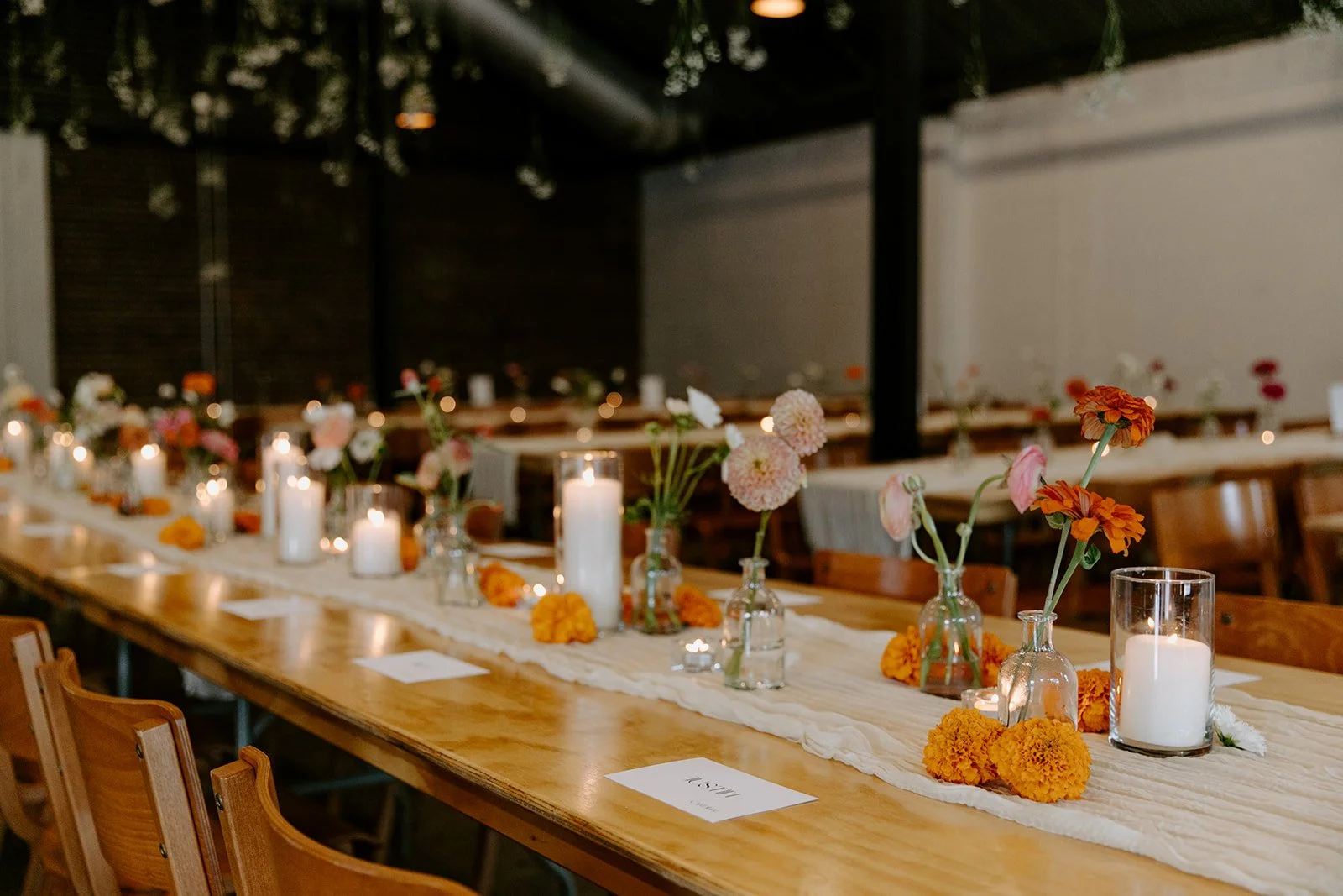 A long wooden table decorated with flowers in small vases, candles, and table cards, set for a celebration or wedding reception in a dimly-lit venue.