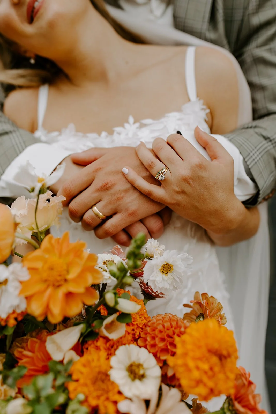 Close-up of a bride and groom's hands with wedding rings, surrounded by orange and white flowers, on her white wedding dress.