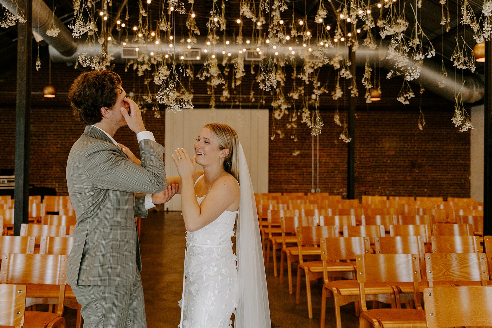 A bride and groom sharing a joyful moment during their wedding ceremony in a decorated indoor venue with hanging flowers and string lights.