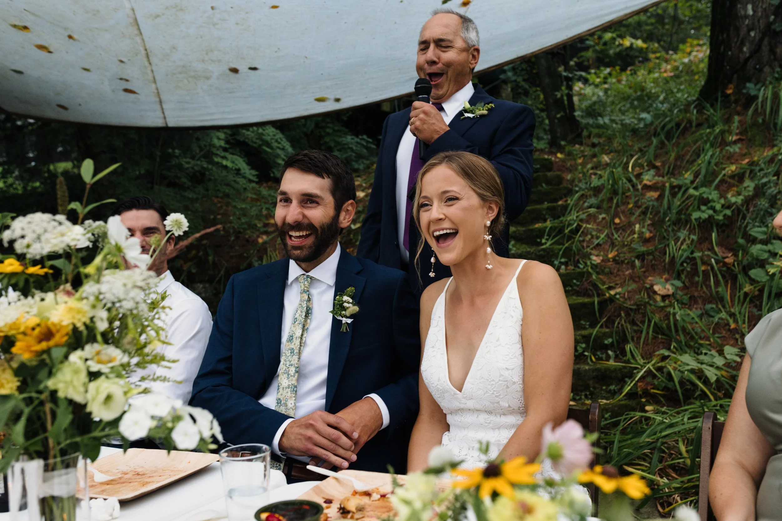 Outdoor wedding reception scene, smiling couple seated at a table, man in a suit giving a toast with a microphone, surrounded by flowers and greenery.