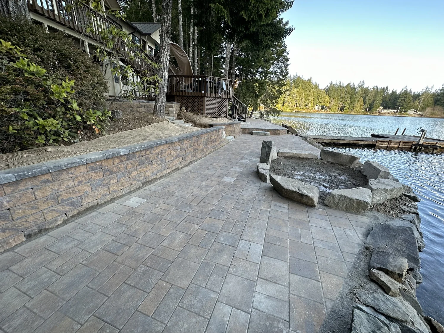 Lakeside paver patio with a curved stone seat wall and stone fire pit at the water's edge, built by Lawn Boys Landscape near Olympia, WA