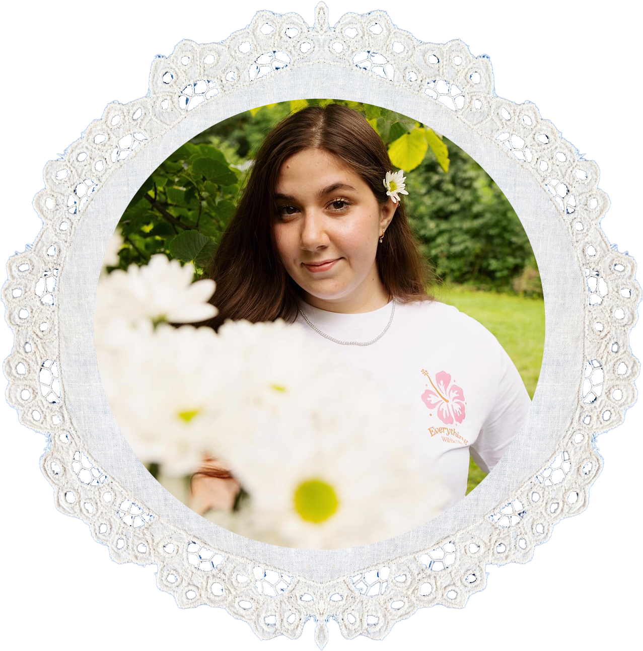 A young woman with long brown hair and a white flower in her hair, standing outdoors among green leaves and trees, smiling slightly at the camera. She is wearing a white shirt with a pink butterfly and the words"Everything" printed on it, and a silver necklace, with a blurred bunch of white flowers in the foreground.