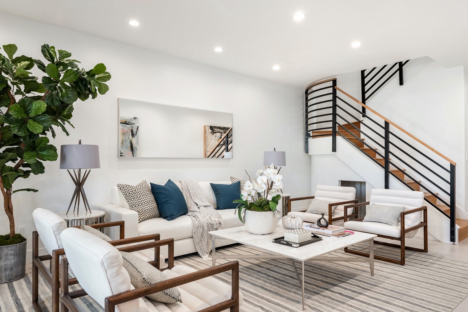 Modern living room with white furniture, indoor plant, abstract art, and a staircase.