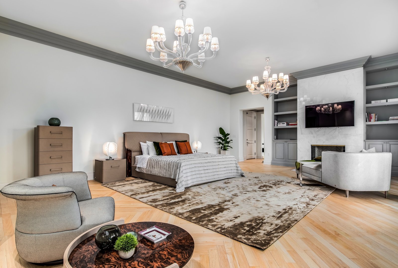 Luxurious modern bedroom with king-size bed, two chandeliers, armchairs, a round coffee table, and built-in shelves. Features neutral tones, hardwood flooring, and sophisticated decor.