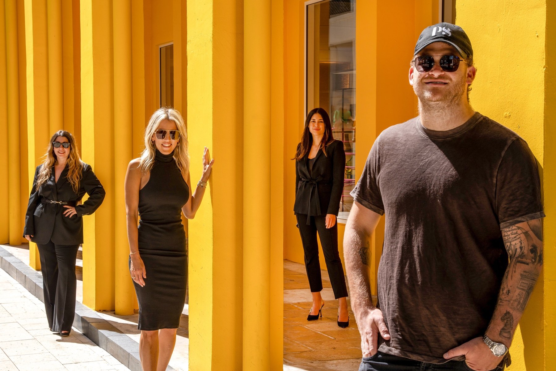 Four people standing in front of yellow columns, three women dressed in black attire and a man in casual clothing with tattoos, wearing sunglasses and a cap.