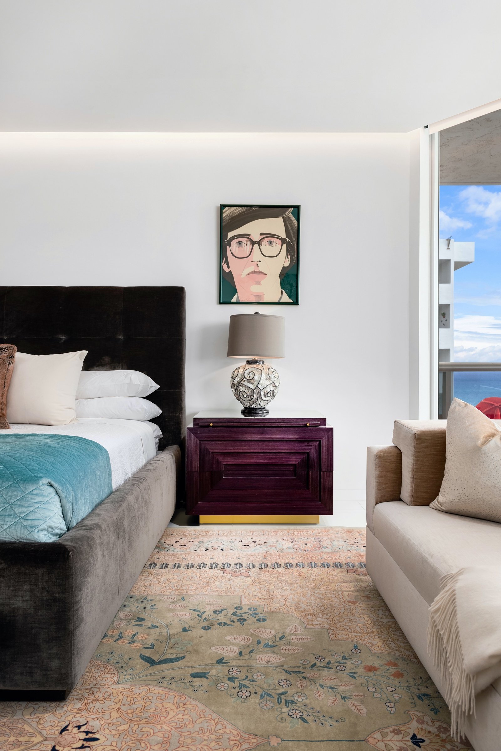 Modern bedroom with a black upholstered bed, teal blanket, and white pillows. Nightstand with a decorative lamp and artwork on the wall above. Large window with a view of the ocean.