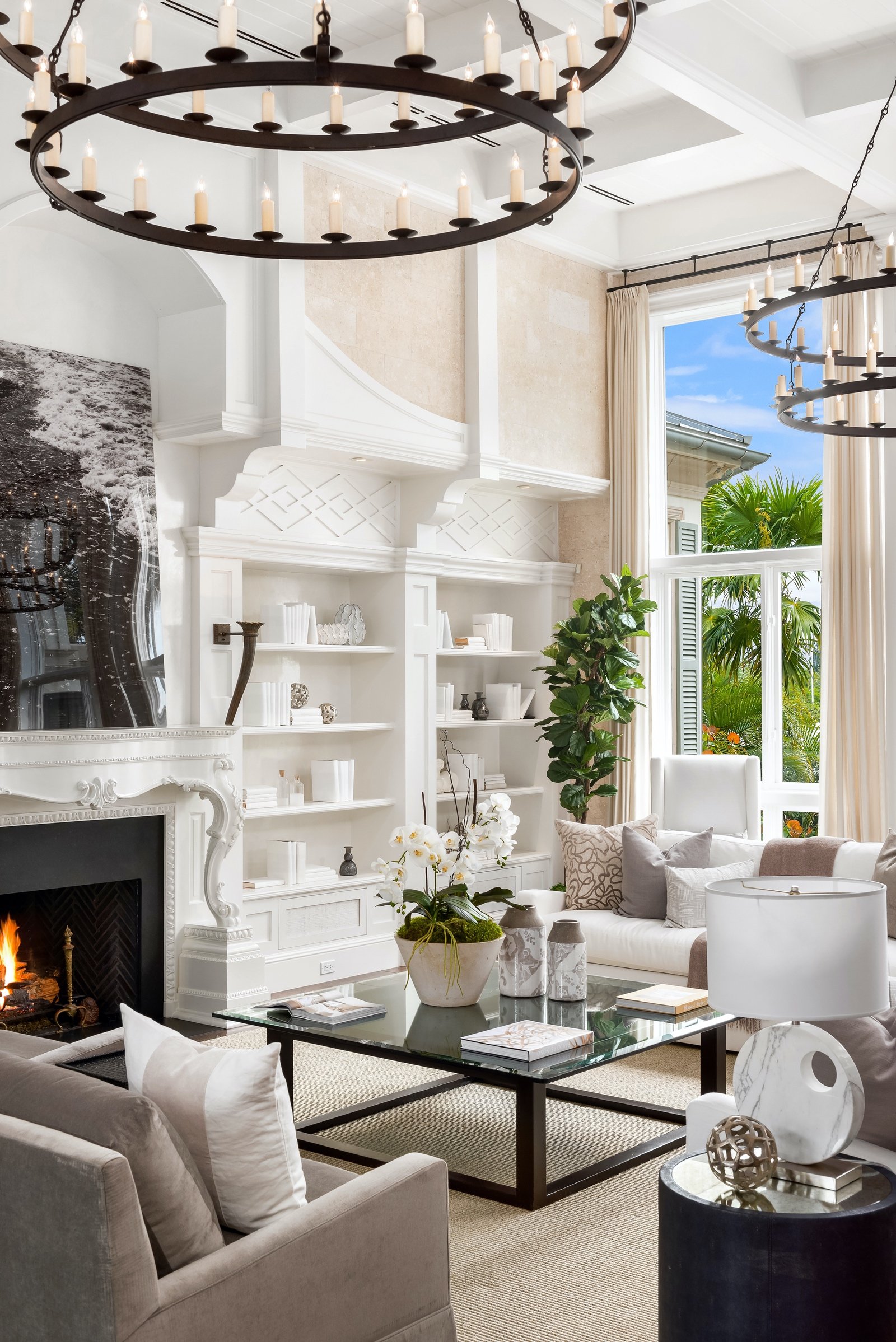 Elegant living room with chandelier, white shelving, fireplace, glass coffee table, potted plant, and large windows with outdoor view. 1 OAK Studios, photo, video, drone, real estate photography and video.