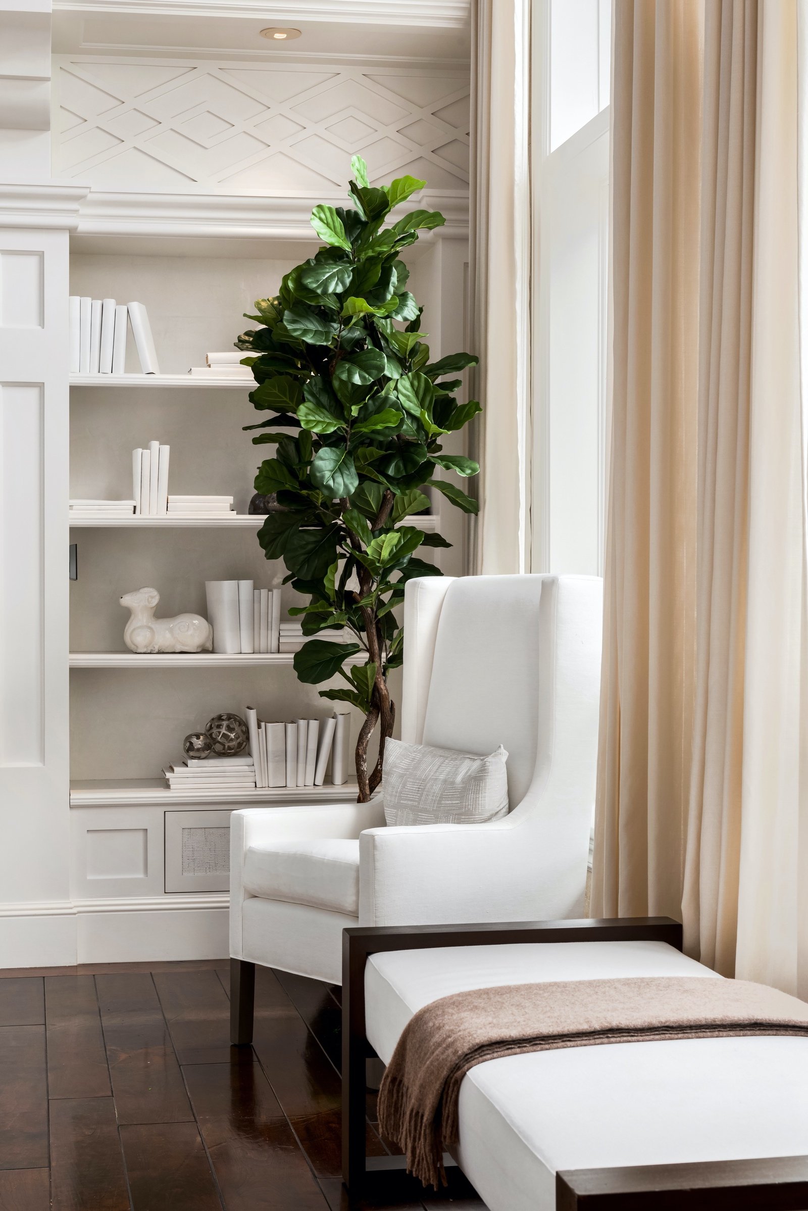 Modern living room with white armchair, indoor plant, beige curtains, bookshelves with white books, and a small sculpture on a dark wood floor.
