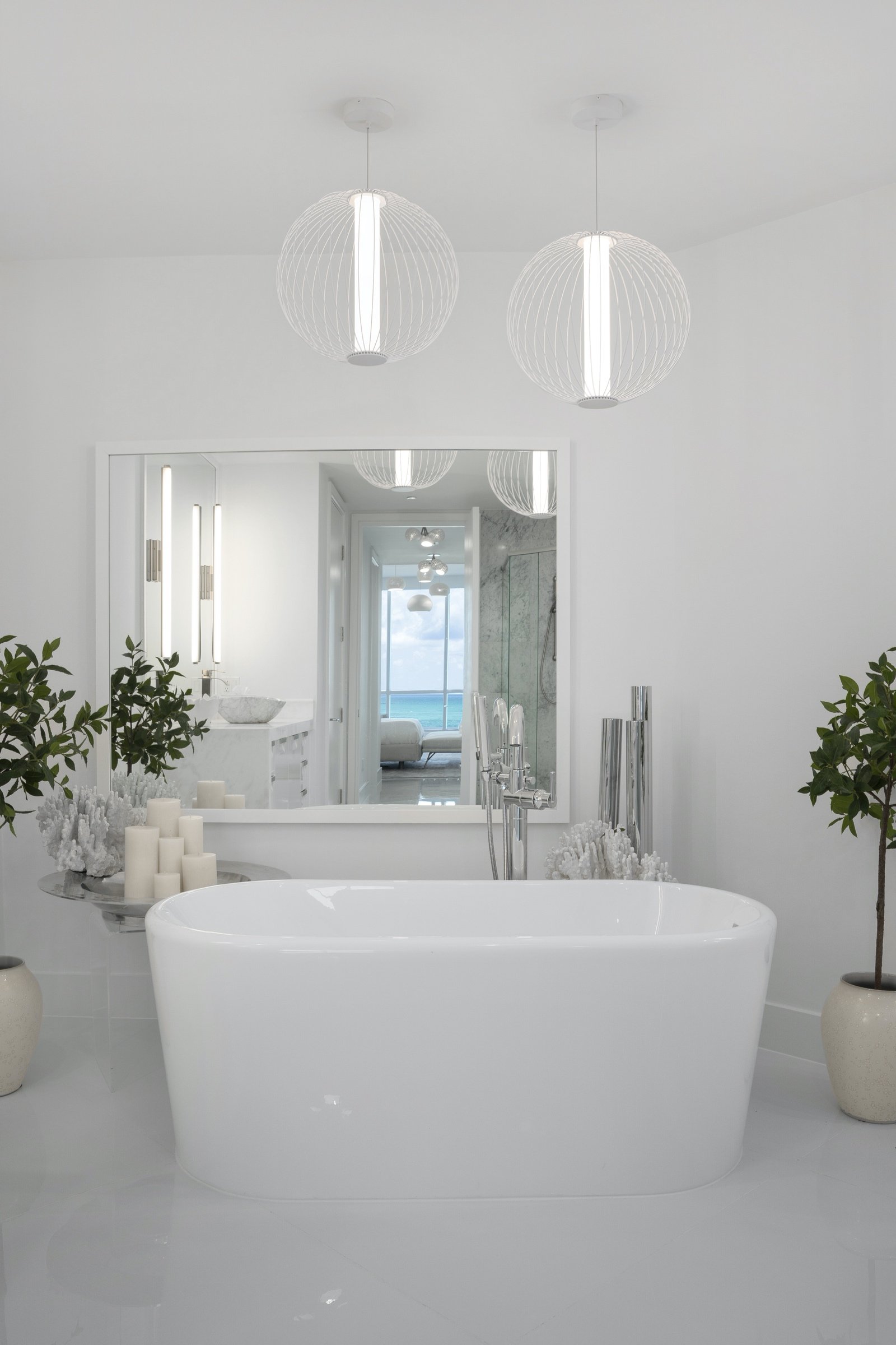 Modern bathroom with freestanding bathtub, large mirror, hanging lights, decorative plants, and ocean view.