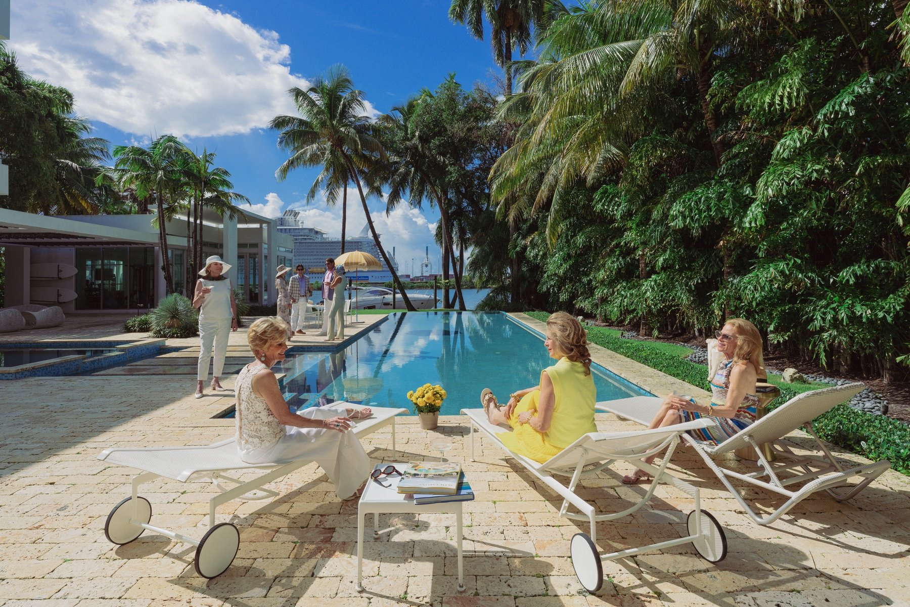 A group of people relaxing by a pool with palm trees and a view of a cruise ship in the background. Several individuals are lounging on chairs while others are standing and chatting. The scene includes tropical plants and a blue sky.