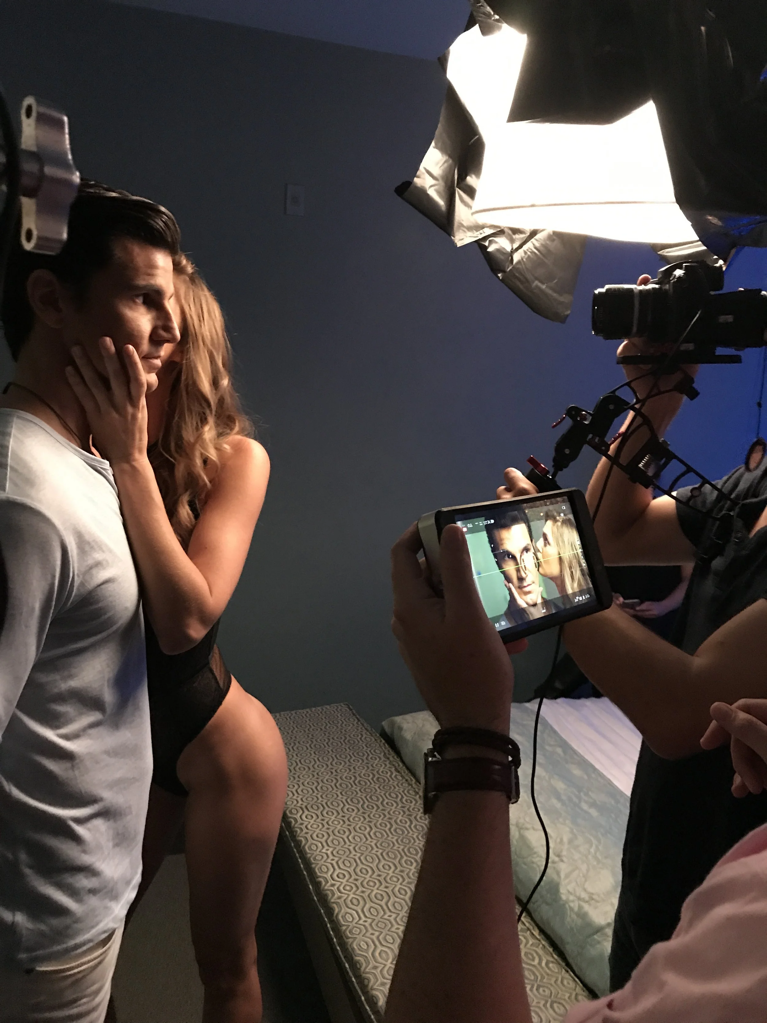 Behind-the-scenes of a photoshoot with a man and woman posing closely, surrounded by cameras and lighting equipment.