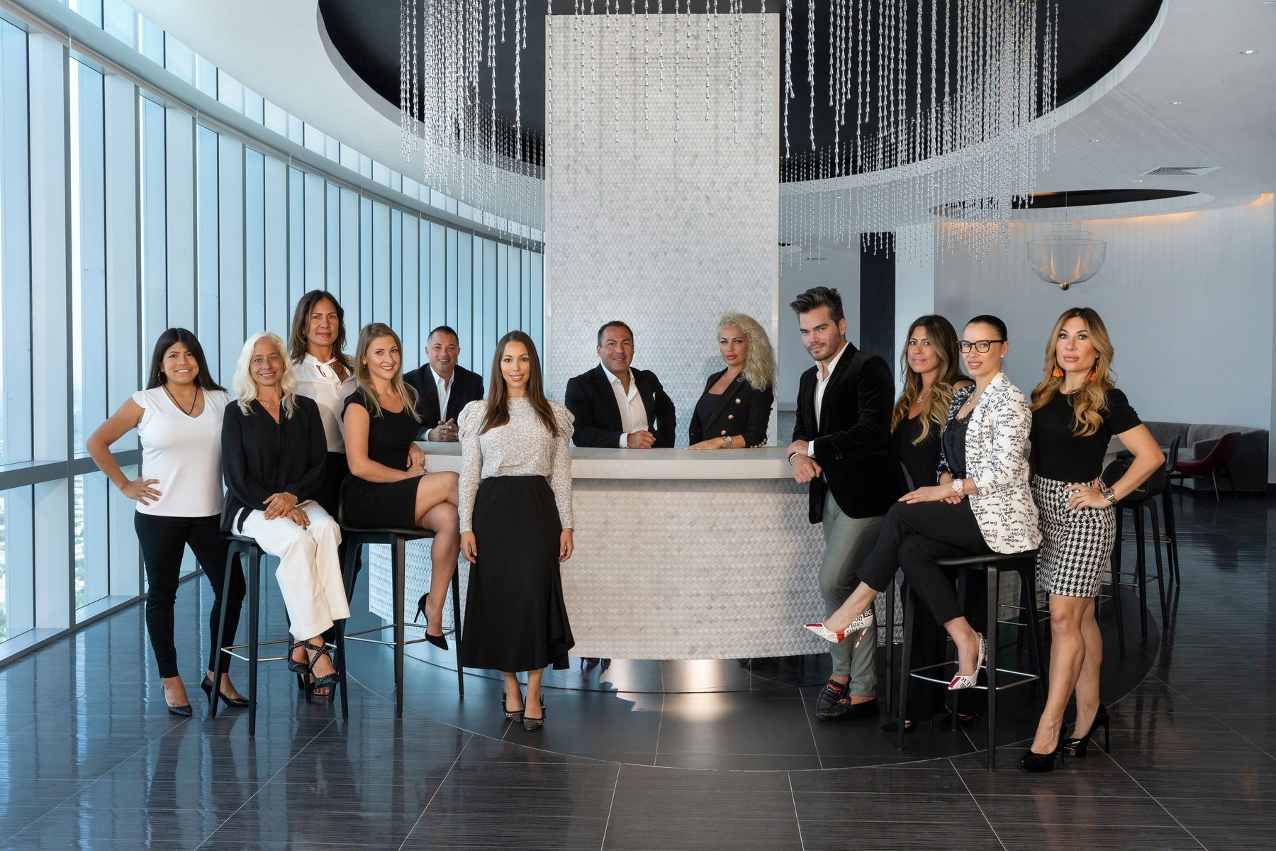 Group of people dressed in formal attire standing and sitting around a bar in a modern office space with floor-to-ceiling windows and decorative hanging lights.