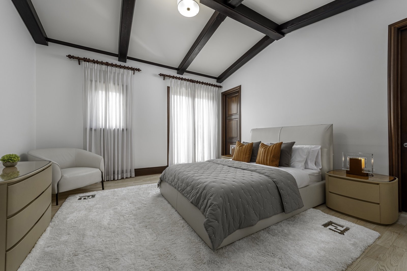 Modern bedroom with a gray quilted bed, white upholstered chair, beige nightstand, and white rug. Two large windows with sheer white curtains and exposed dark wooden ceiling beams.