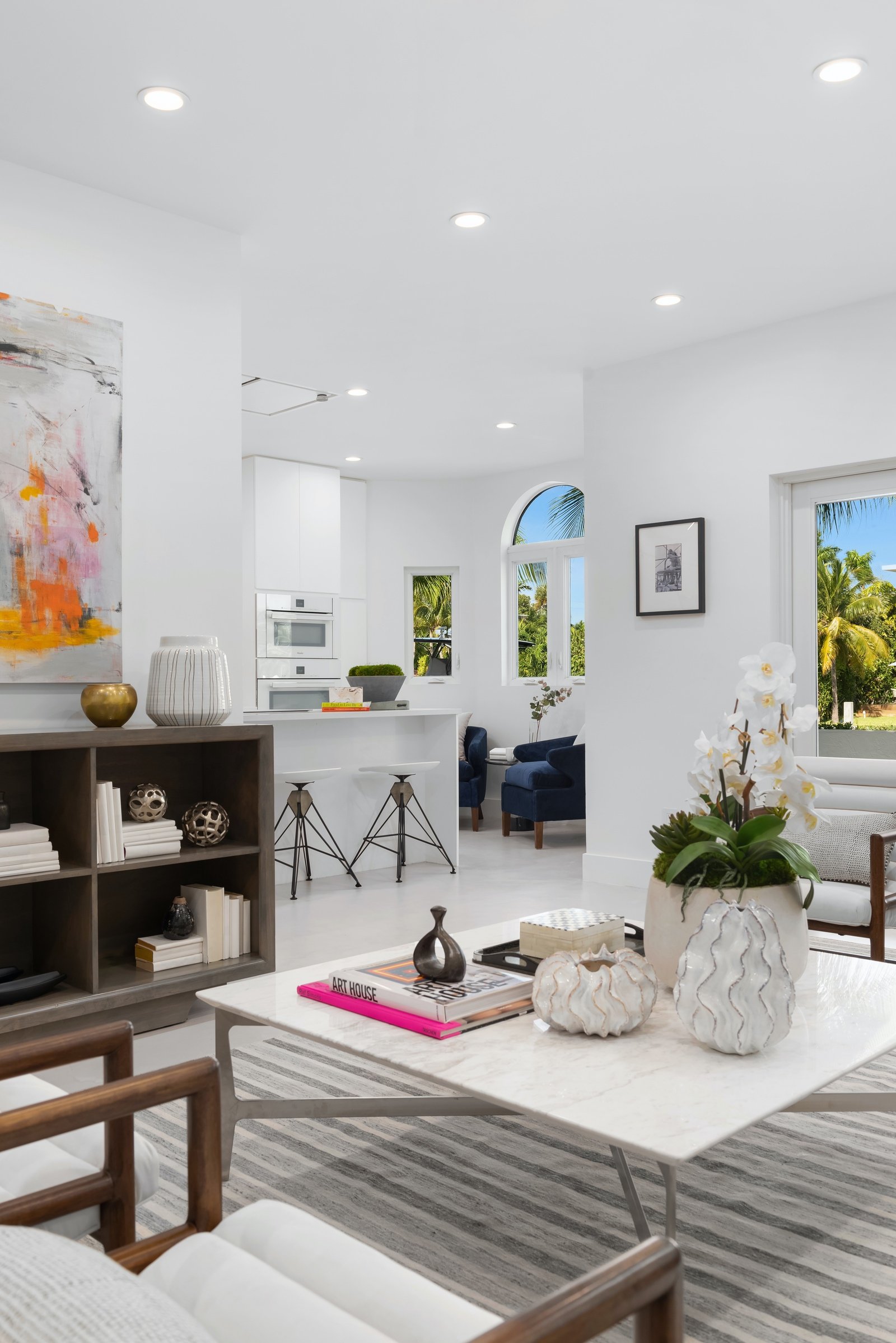 Modern living room and kitchen with white walls, abstract painting, indoor plants, minimalist furniture, and decorative items on shelves and table.