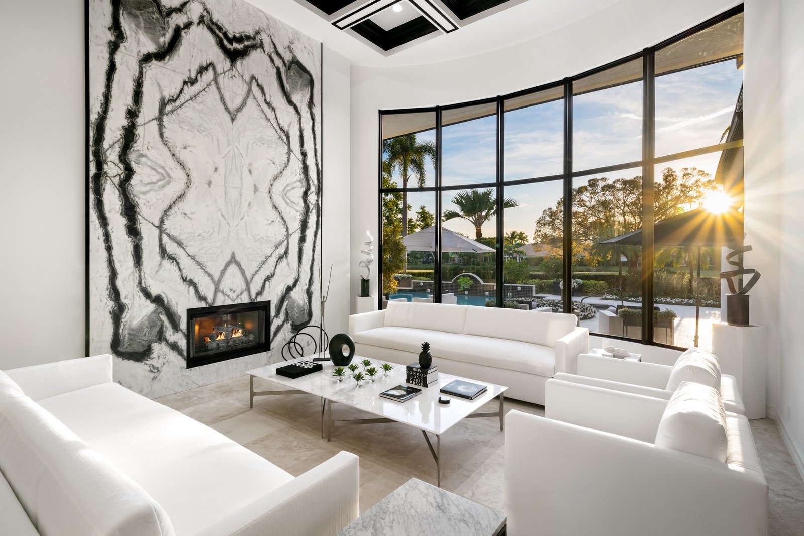 Modern living room with white sofas, marble fireplace, large windows displaying garden view and sunset, contemporary decor."}