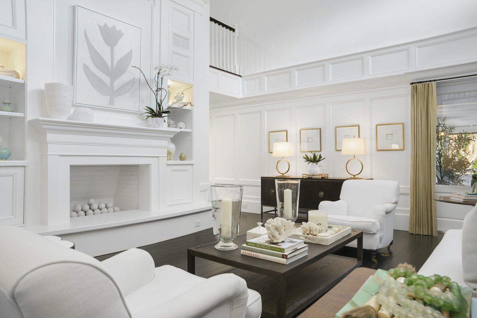 Modern white living room with a fireplace, white furniture, a coffee table with books and decorative items, and a console table with lamps. Shelves are built into the wall next to the fireplace. The room has a neutral palette with accent lighting and