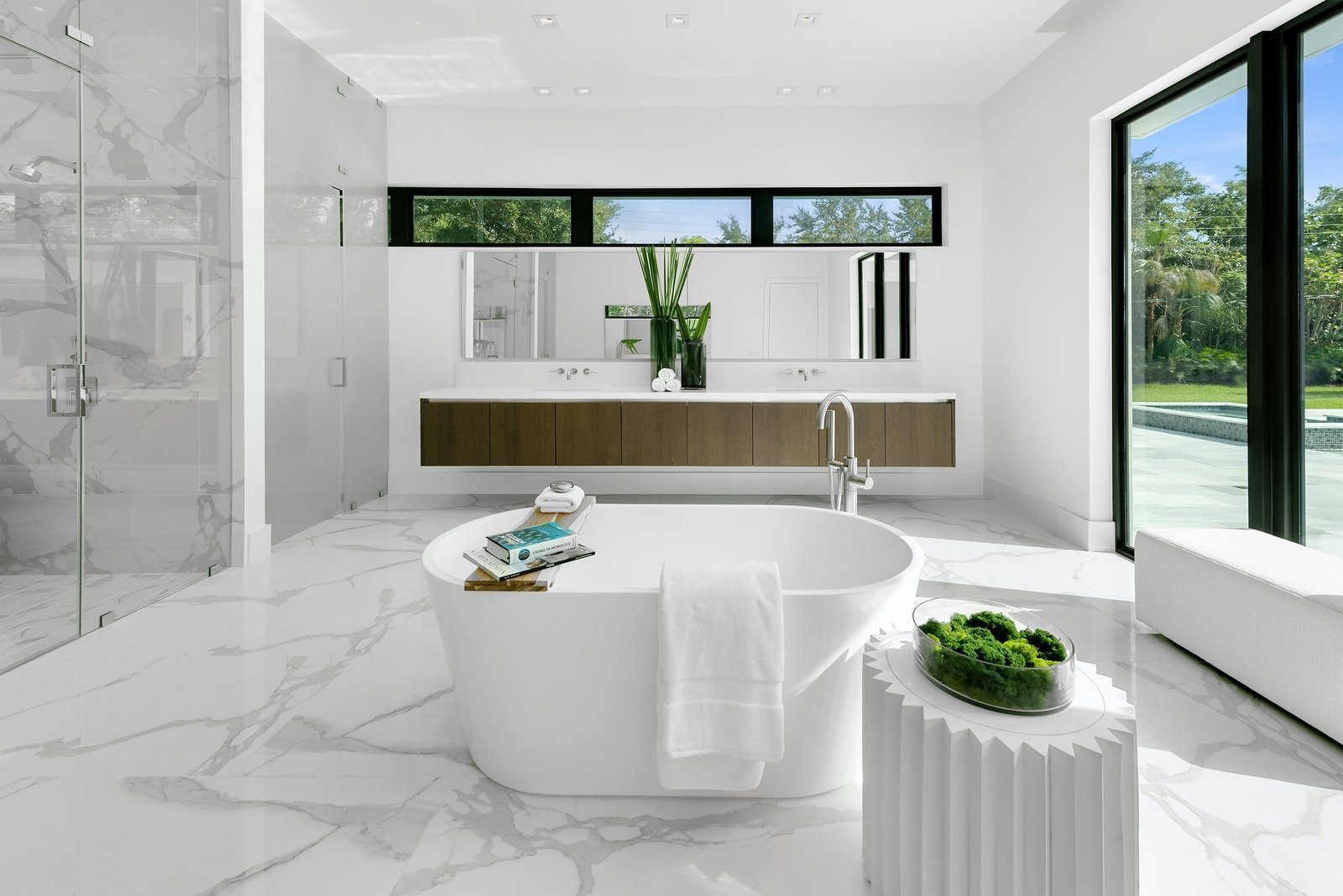 Modern bathroom with freestanding tub, white marble flooring, large mirrors, floating vanity, and a window view of garden.