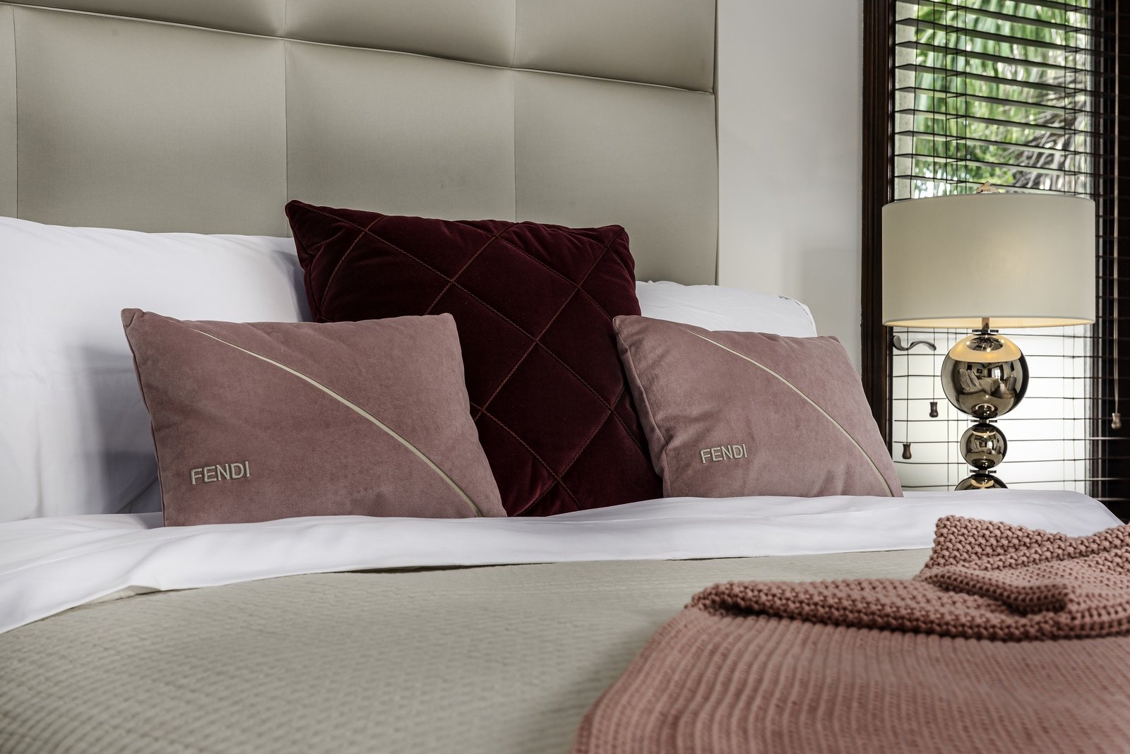 Luxurious bedroom with beige headboard, pillows marked with 'Fendi,' a knit throw blanket, and a modern lamp by the window.