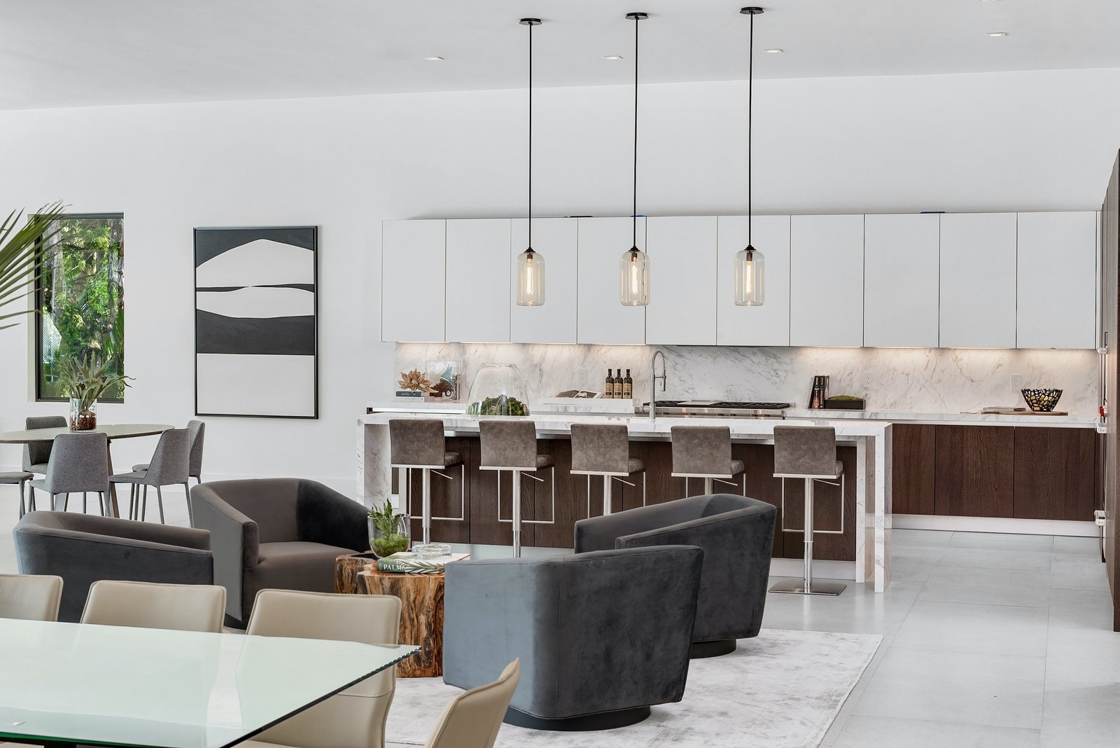 Modern open-plan living area featuring a kitchen with marble countertops, pendant lighting, bar stools, and white cabinetry. A dining table with chairs, and contemporary lounge chairs around a small table are also visible. A large abstract painting a