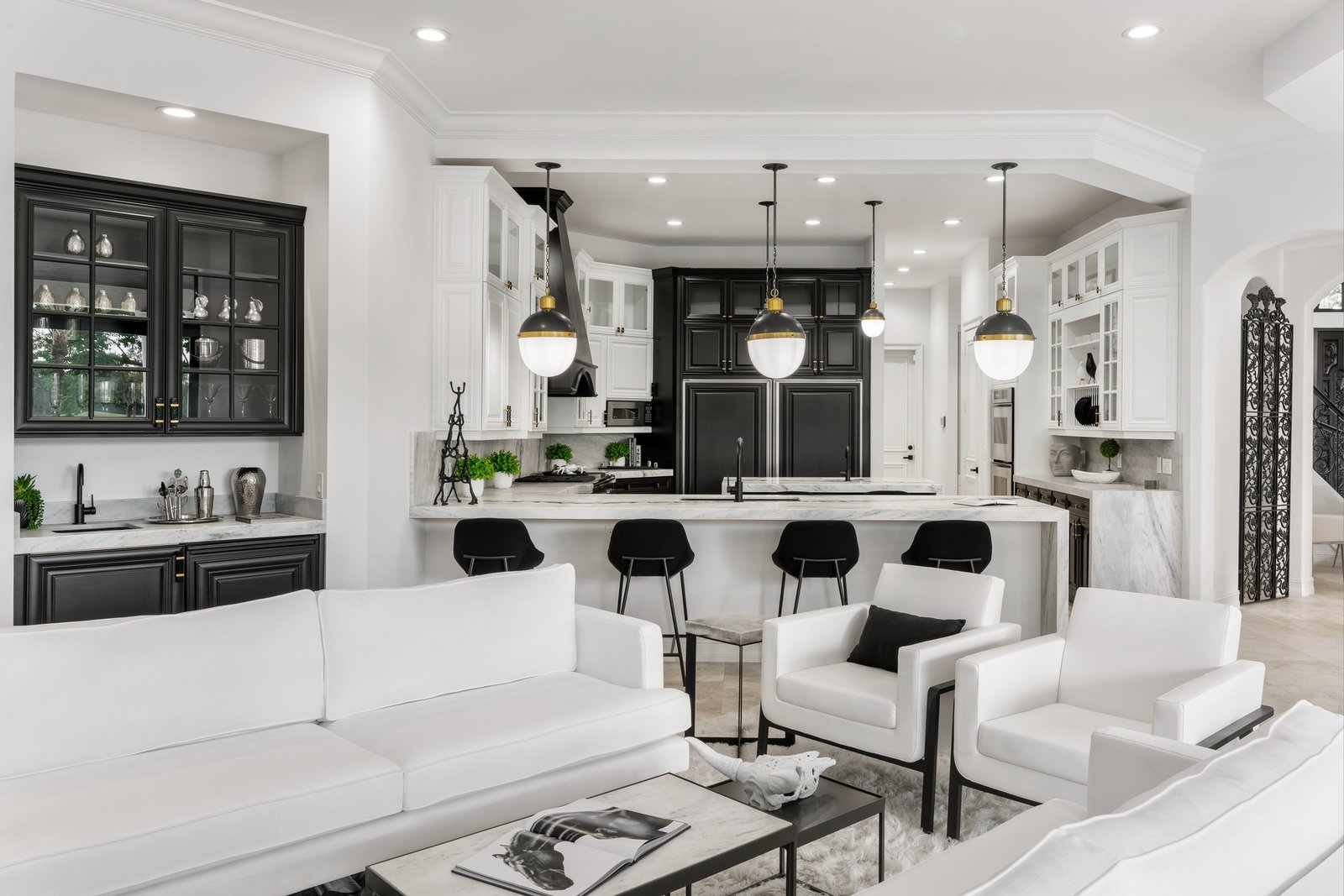 Modern open kitchen and living room with white and black decor, pendant lighting, black cabinets, white sofas and chairs, marble countertops, and bar stools.