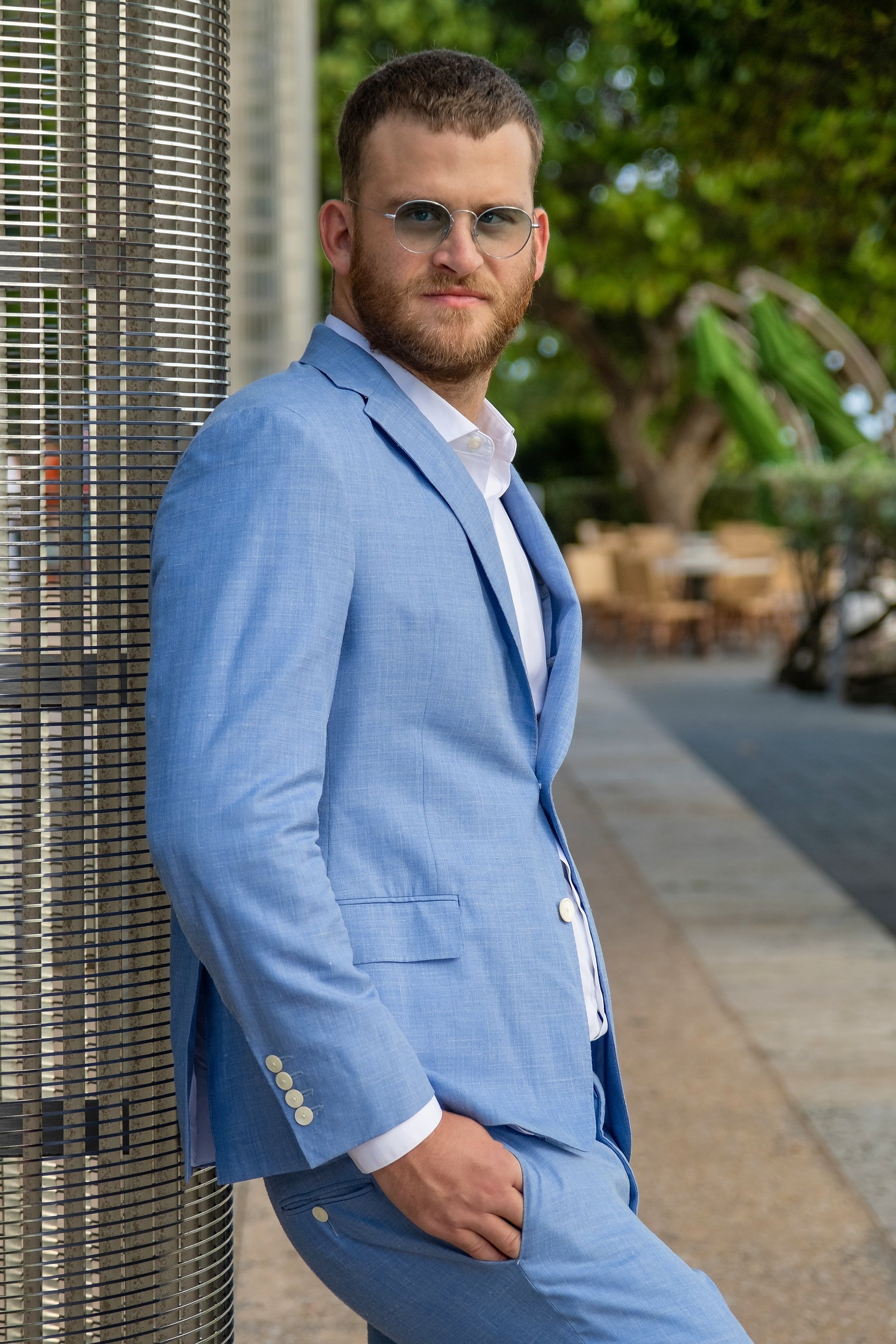 Man in a light blue suit leaning against a metal grid with an outdoor setting in the background.