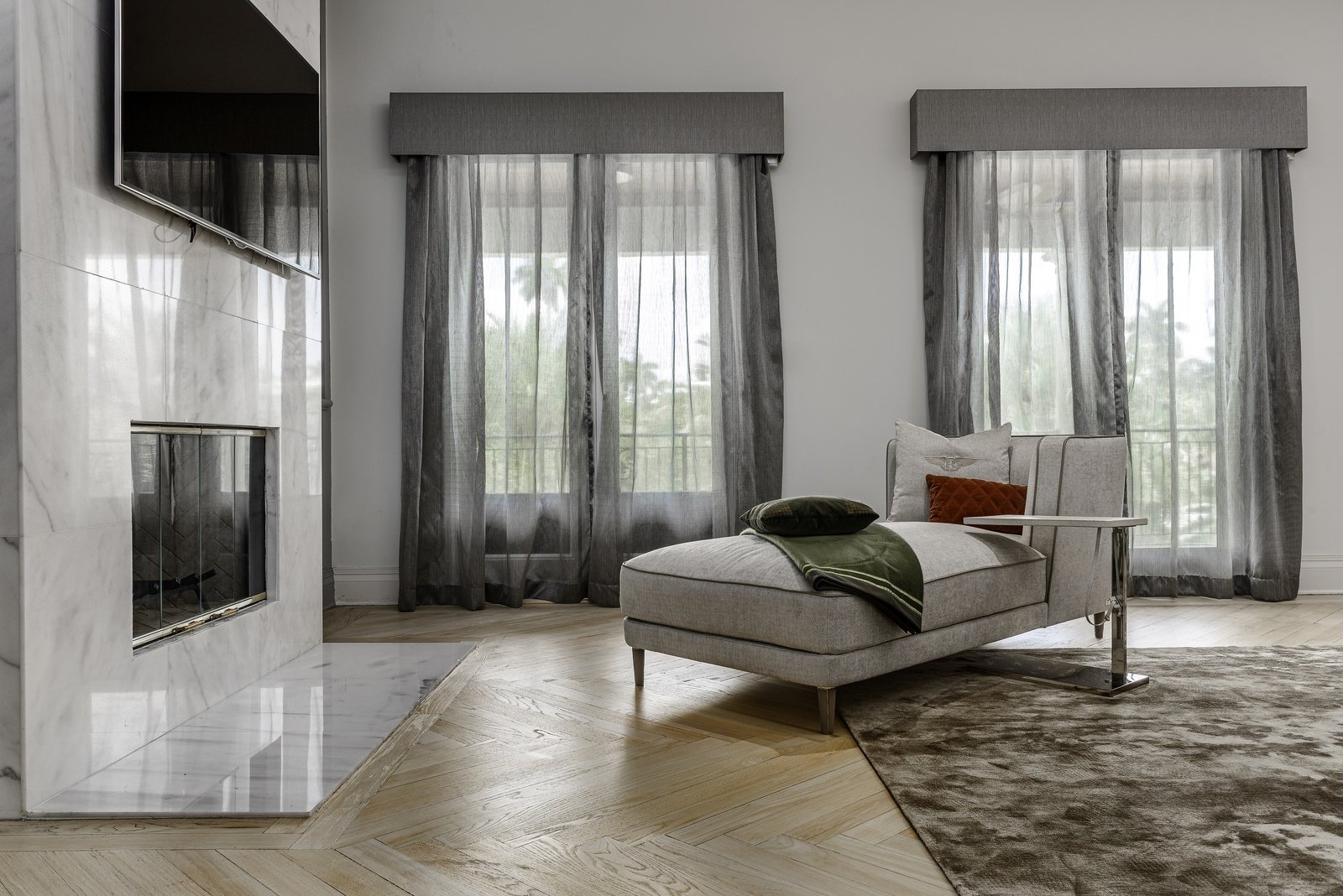 Elegant living room with a chaise lounge, marble fireplace, large windows with sheer curtains, and a wall-mounted TV.