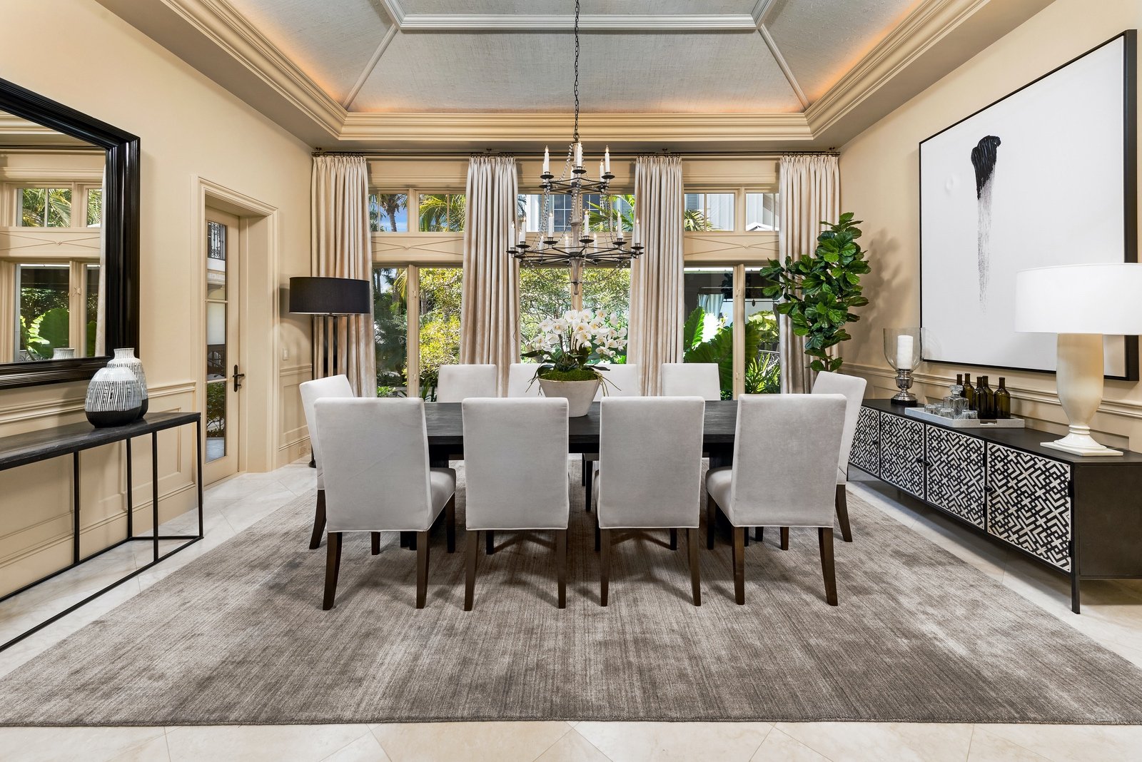 Elegant dining room with a large rectangular table and eight upholstered chairs. The room features a chandelier, large windows with curtains, a sideboard with decorative items, a floor lamp, and a modern painting. There's a large mirror and plant, an