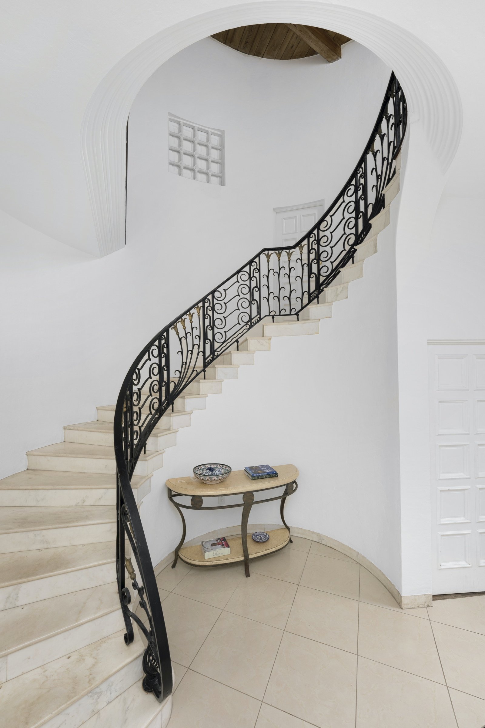 Elegant curved staircase with black wrought iron railing and beige stone steps, leading to an upper floor. Below is a small decorative table with books and a ceramic dish, set against a white wall with a curved design.