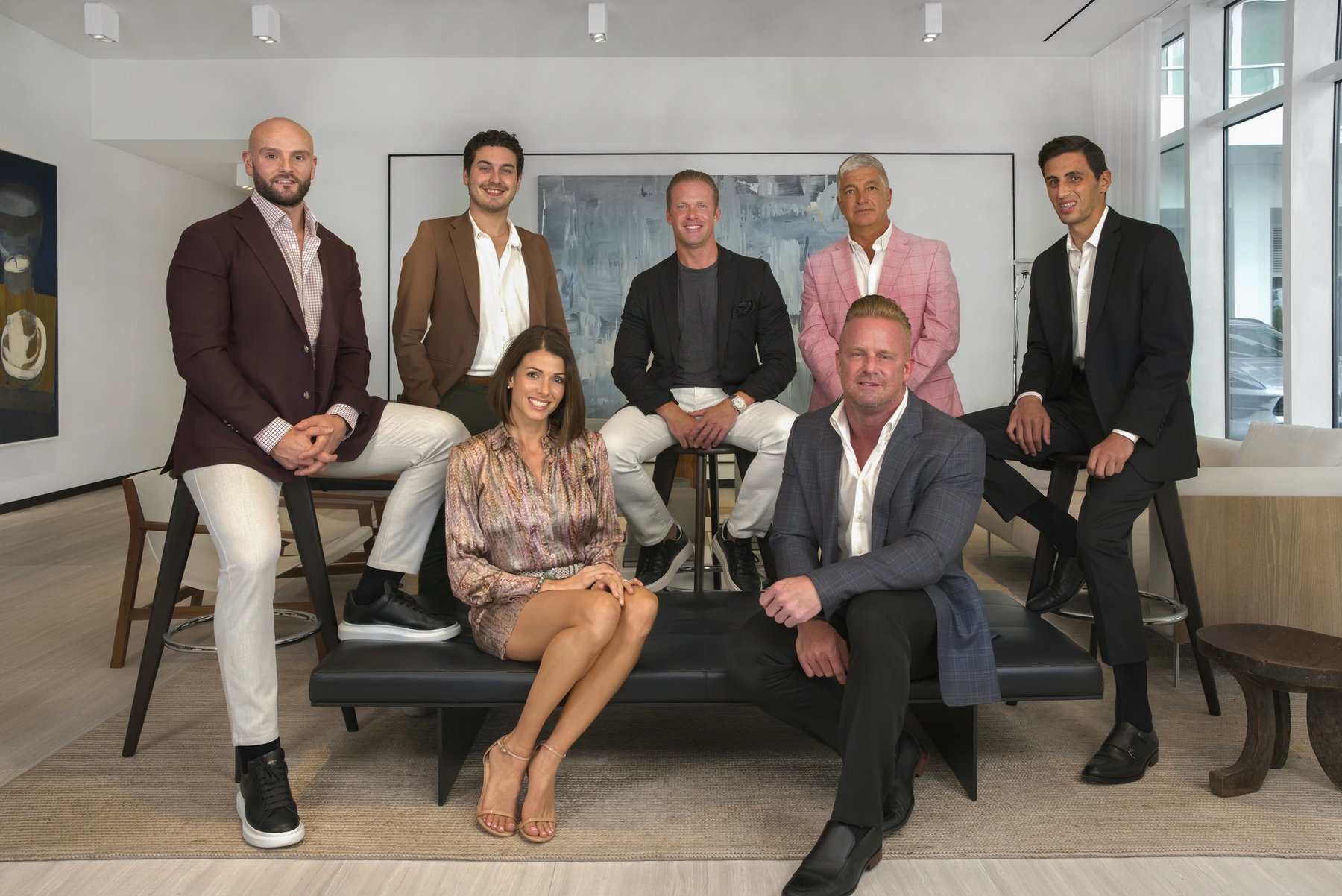 Group of six men and one woman in business attire sitting and standing in a modern office setting.