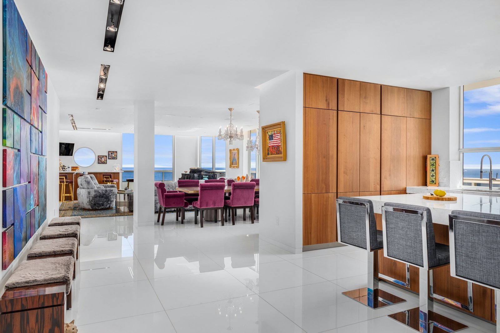 Modern open-concept living space with vibrant decor, including colorful wall art, purple dining chairs, gray bar stools, and wooden cabinetry. Large windows provide an ocean view.