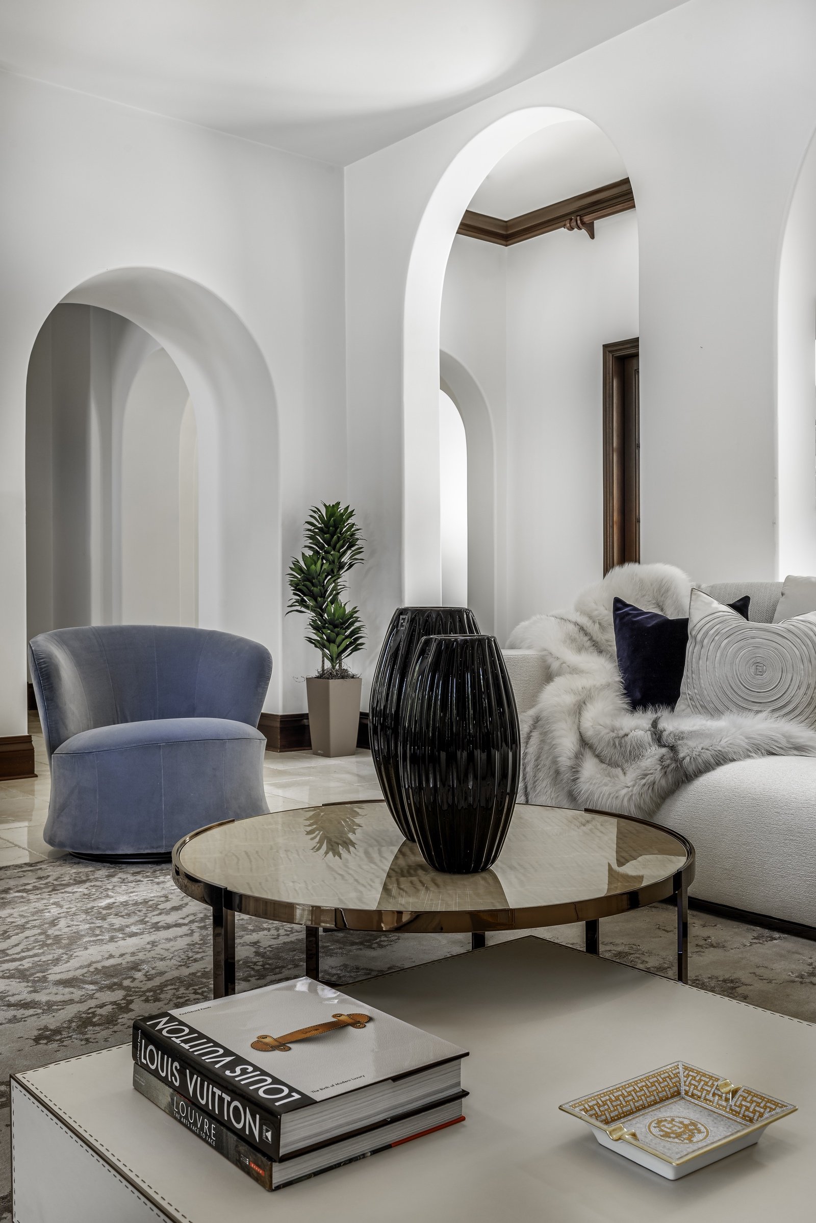 Modern living room with arched white walls, a blue chair, a round glass coffee table with a black vase, a white sofa with pillows and a throw blanket, a potted plant, and books on a side table.