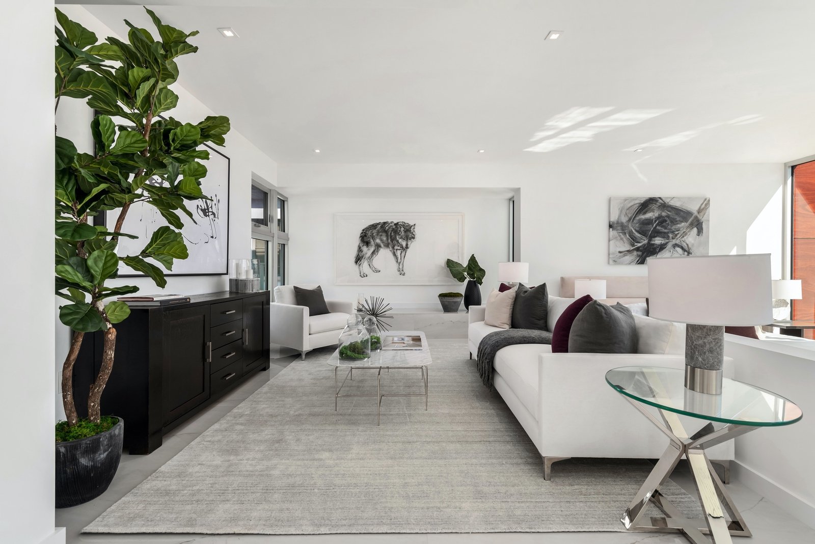 Modern living room with white sofas, glass coffee table, and large indoor plant. Black sideboard with abstract art on the wall. Light gray rug and large windows allow natural light. Minimalist decor.