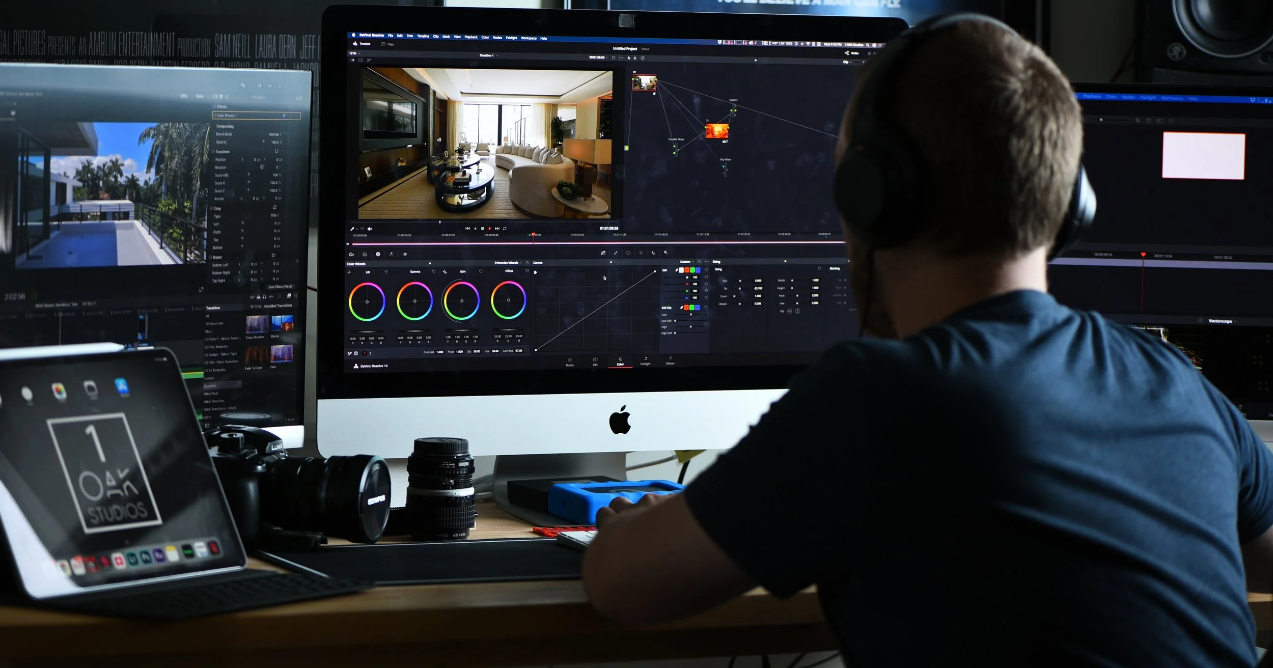 Person editing video on a desktop computer with multiple monitors in a home studio, featuring color grading software and camera equipment on the desk.