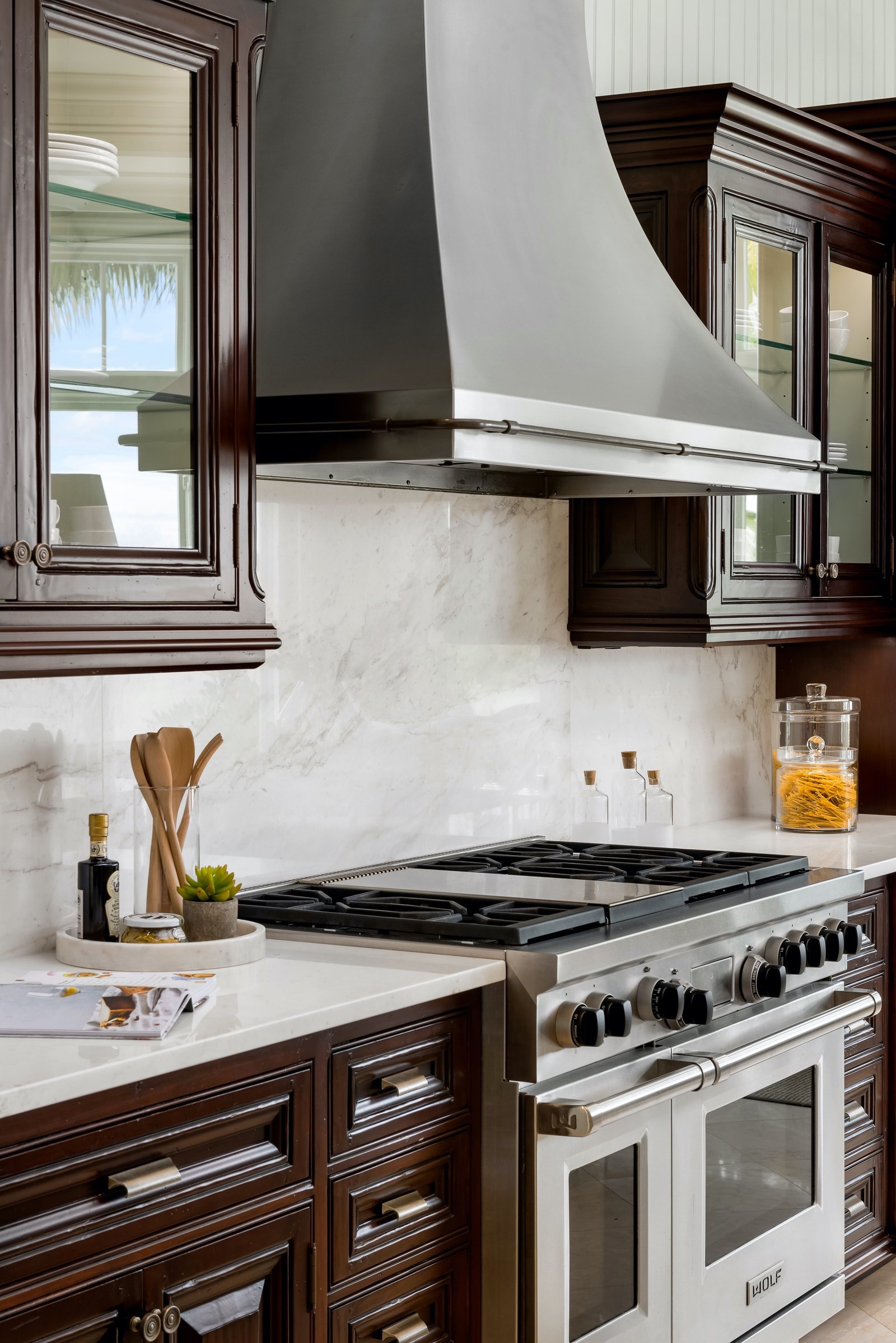 Modern kitchen with stainless steel stove and range hood, marble backsplash, dark wood cabinets, and decor items including potted plant and glass jars.