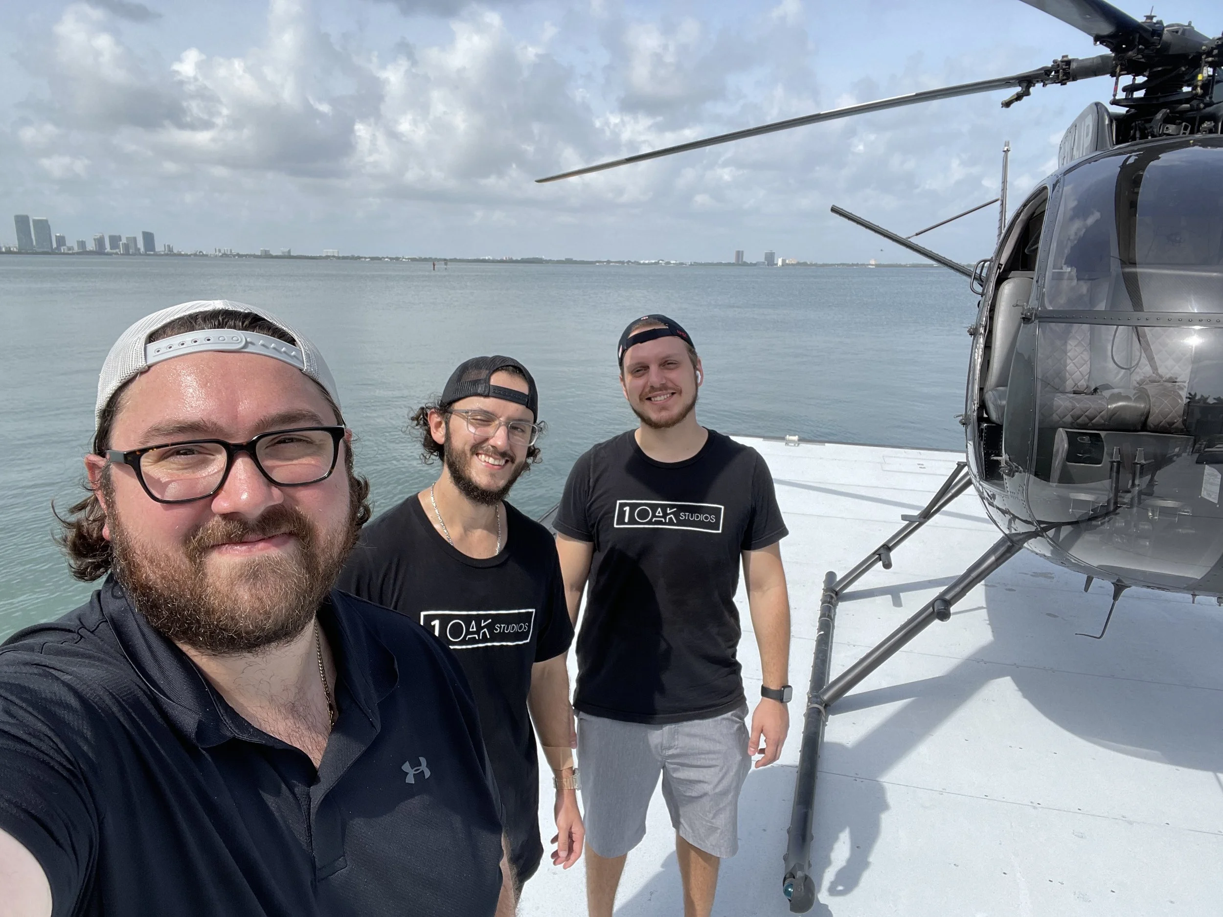 Three men standing on a helipad next to a helicopter with water and a city skyline in the background. 1 OAK Studios, photo, video, drone, real estate photography and video.