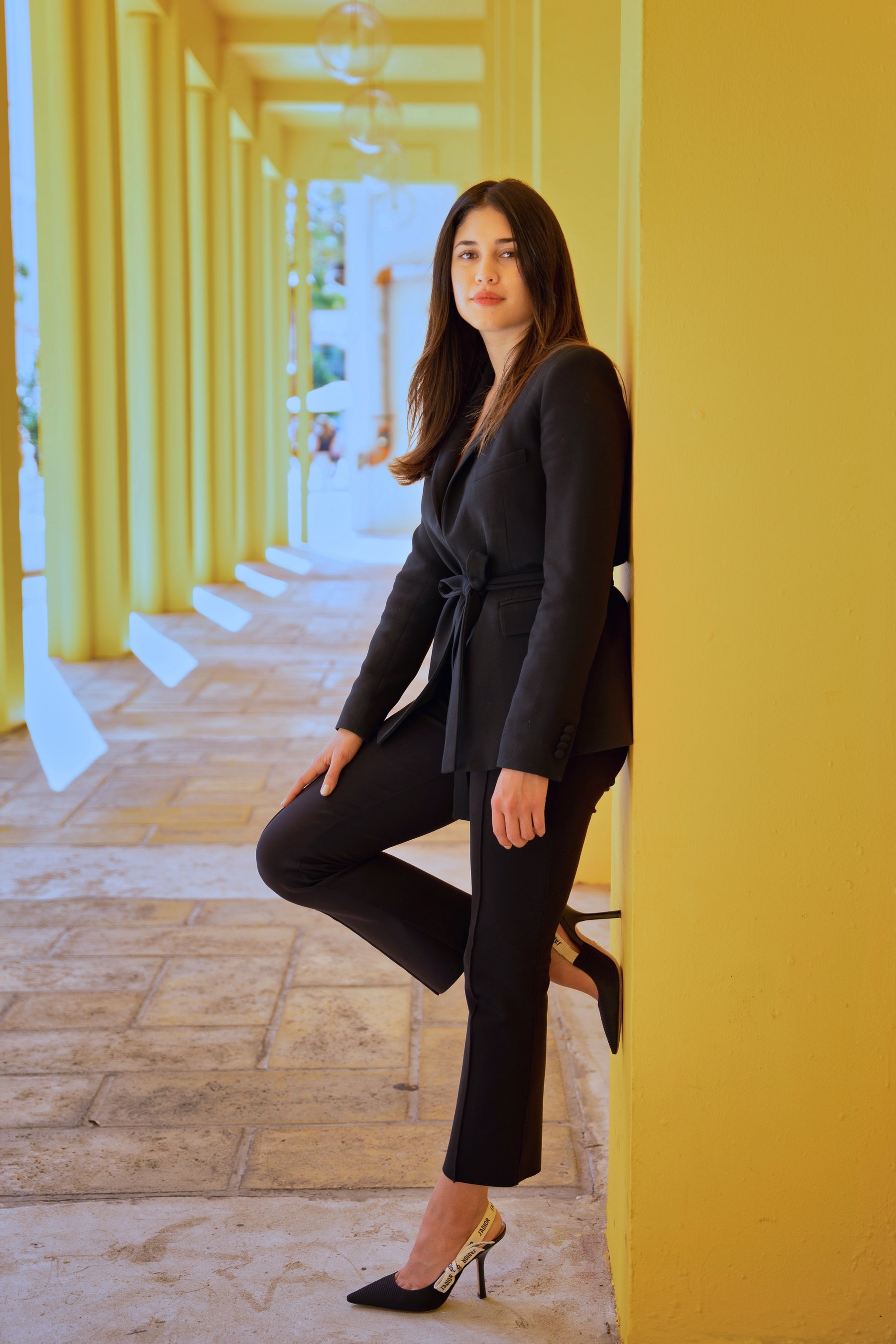 Woman in black outfit leaning against a yellow wall in a corridor with natural lighting.