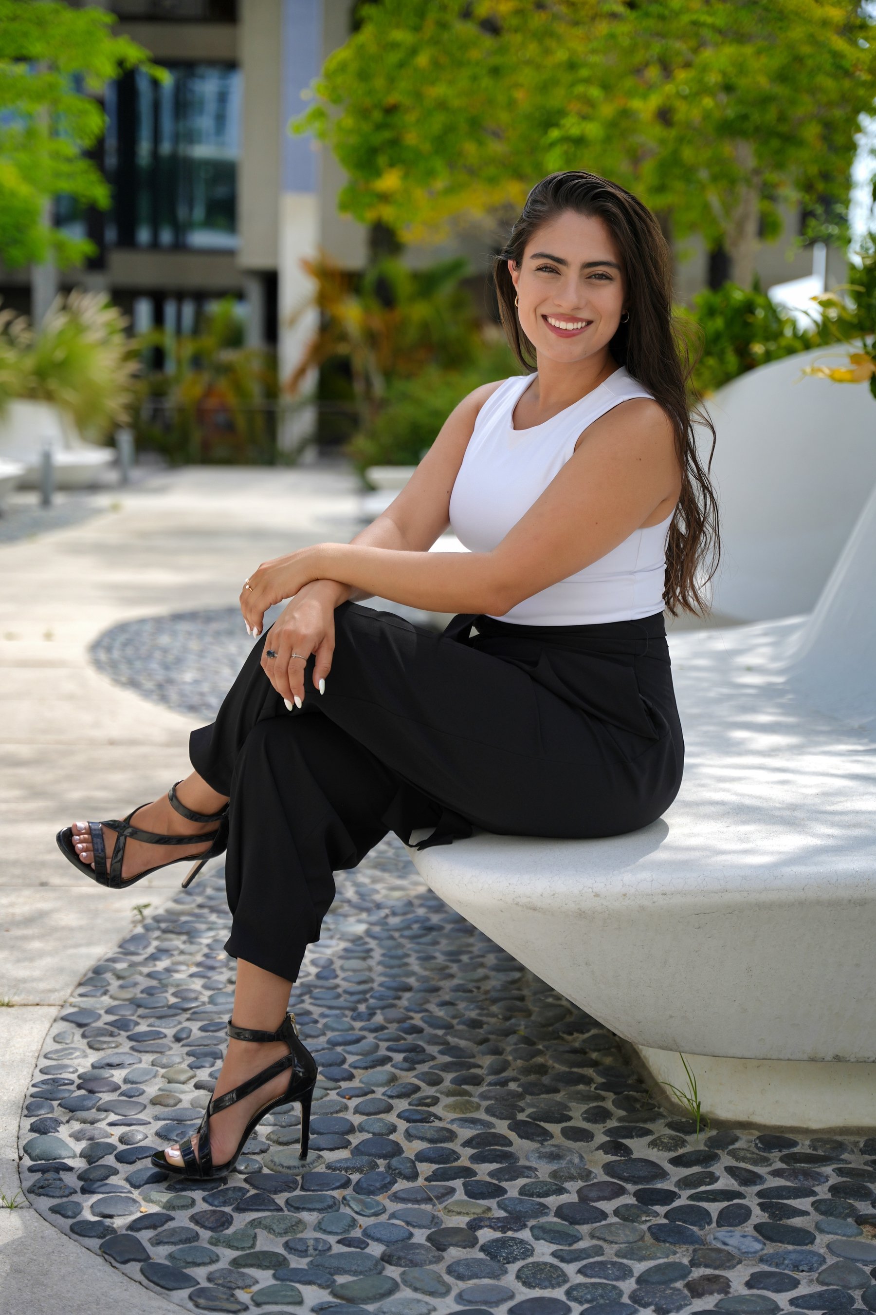 Woman in white top and black pants sitting on an outdoor bench, smiling.