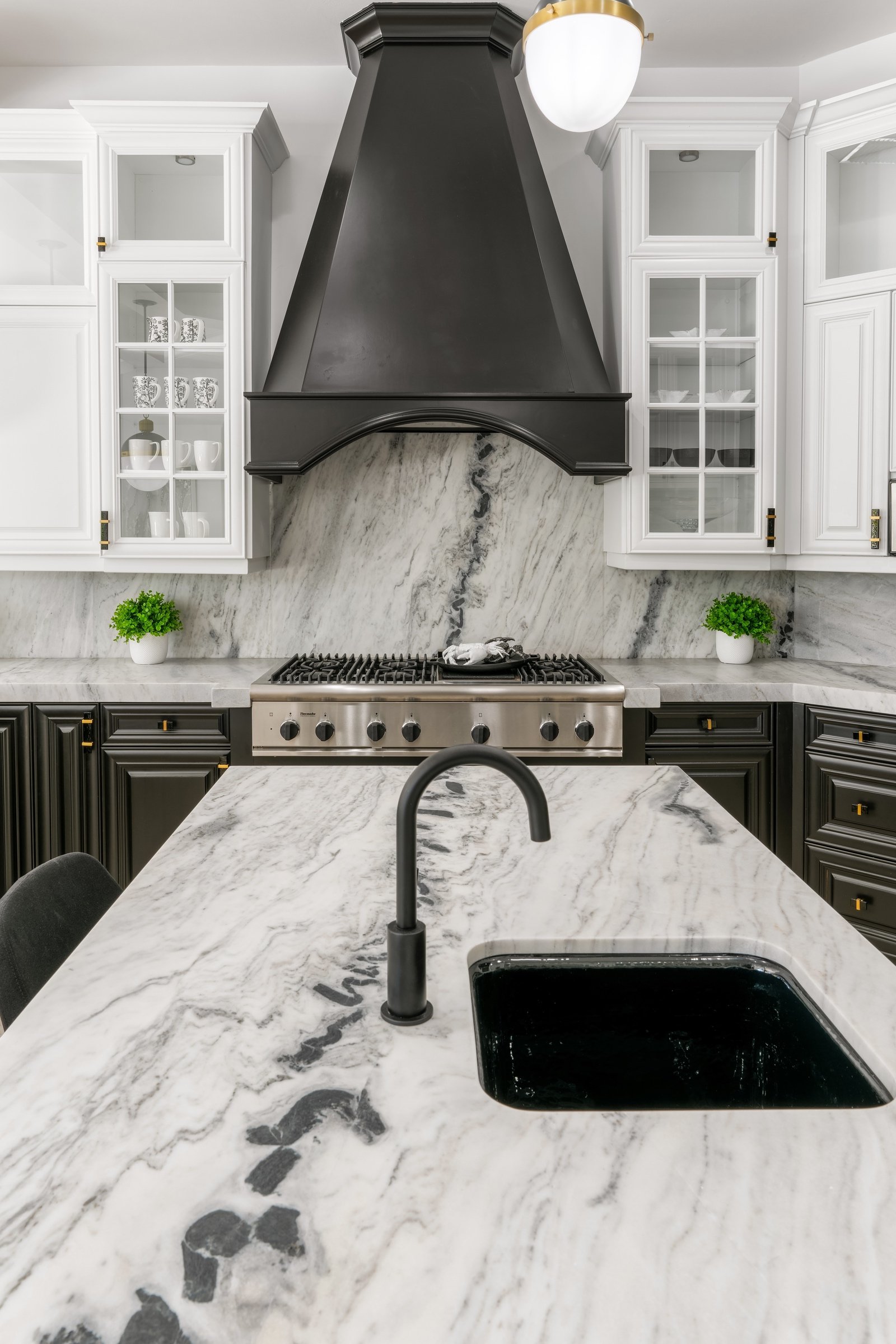 Modern kitchen with marble countertops, black range hood, glass cabinets, stainless steel stove, and black faucet.