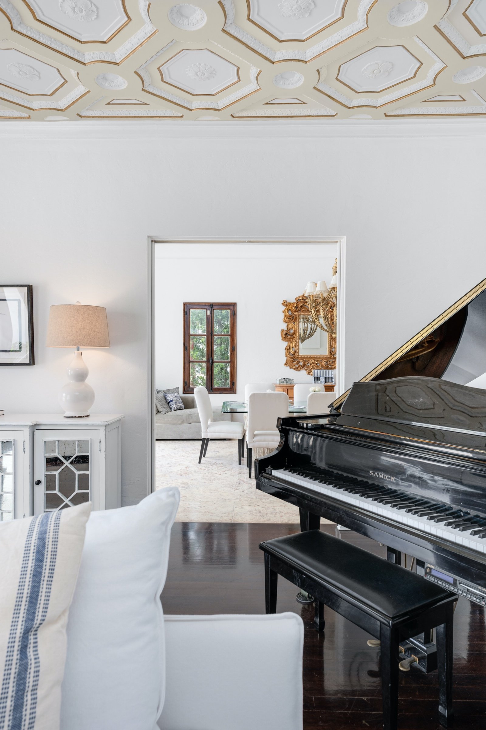Elegant living room with a black grand piano, decorative ceiling, white furniture, and ornate mirror.