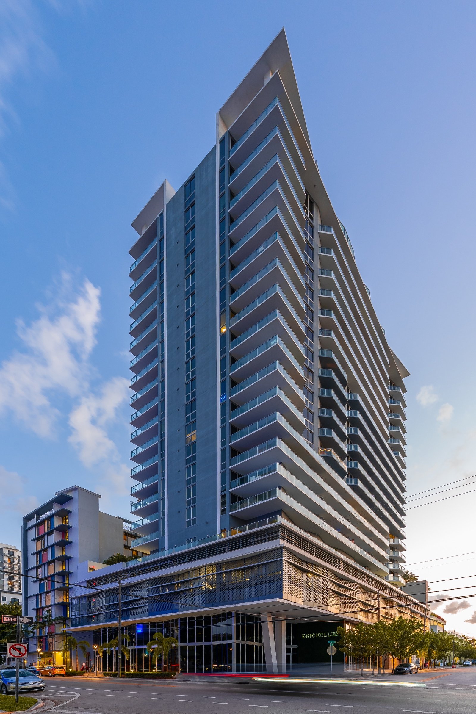 Tall modern apartment building with glass balconies, set against a clear blue sky.