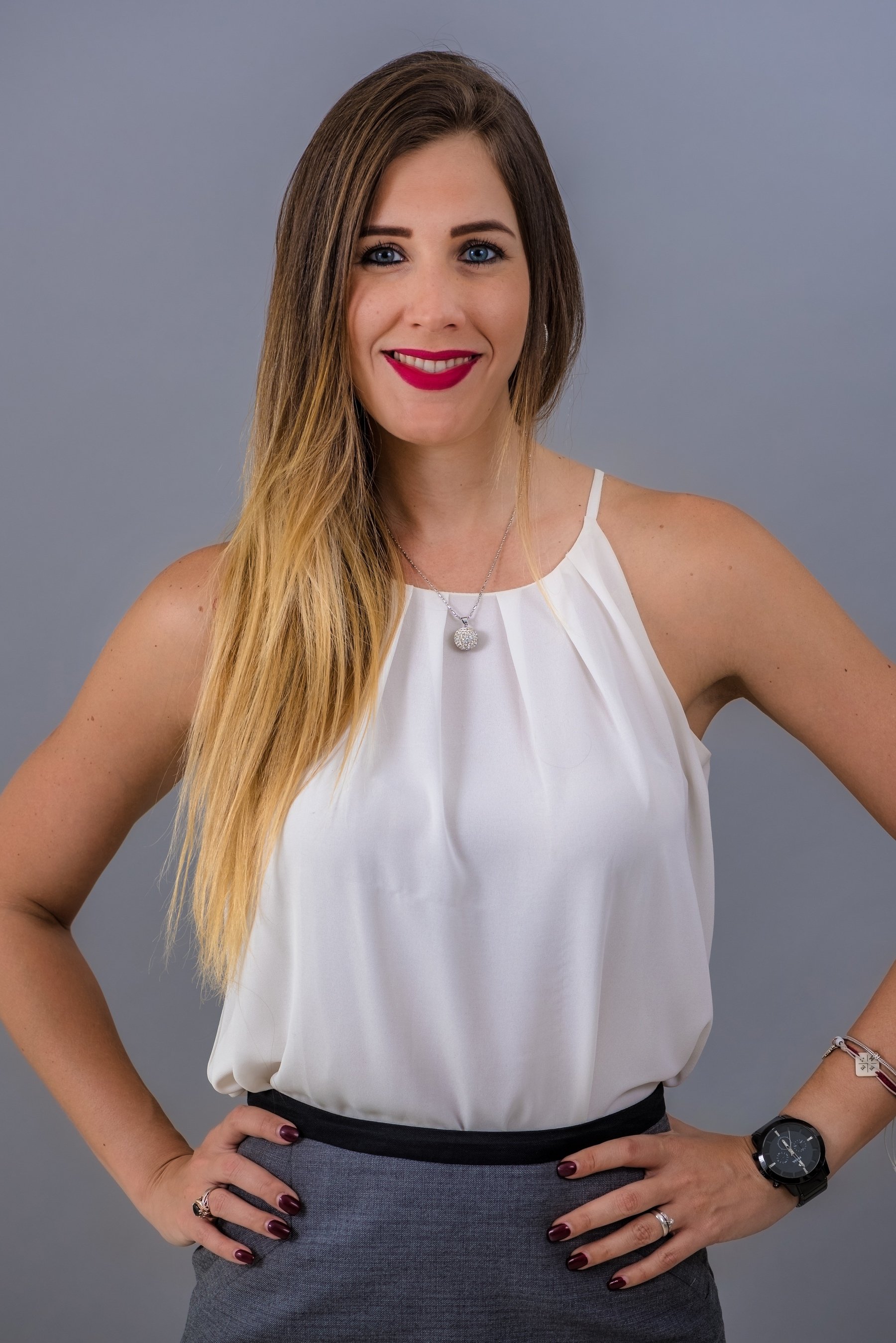 Woman with long hair and bright lipstick wearing a white sleeveless top and gray pants, posing with hands on hips, against a gray background.