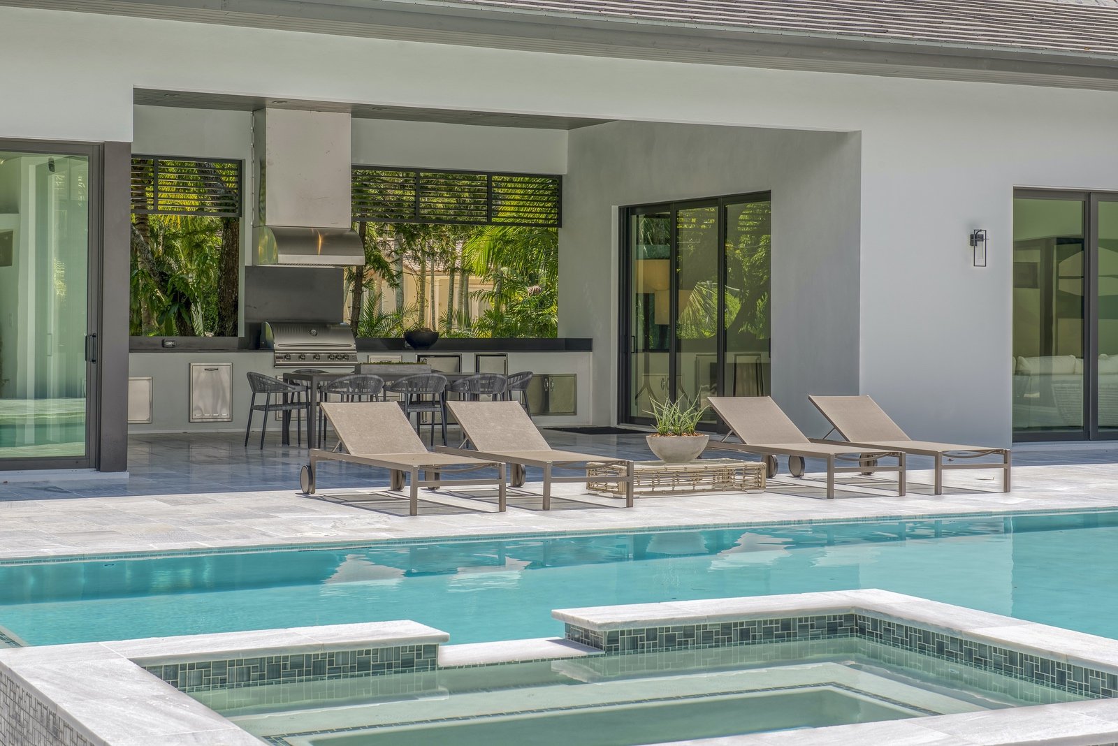 Modern outdoor patio with lounge chairs, swimming pool, and covered dining area with a grill and table.