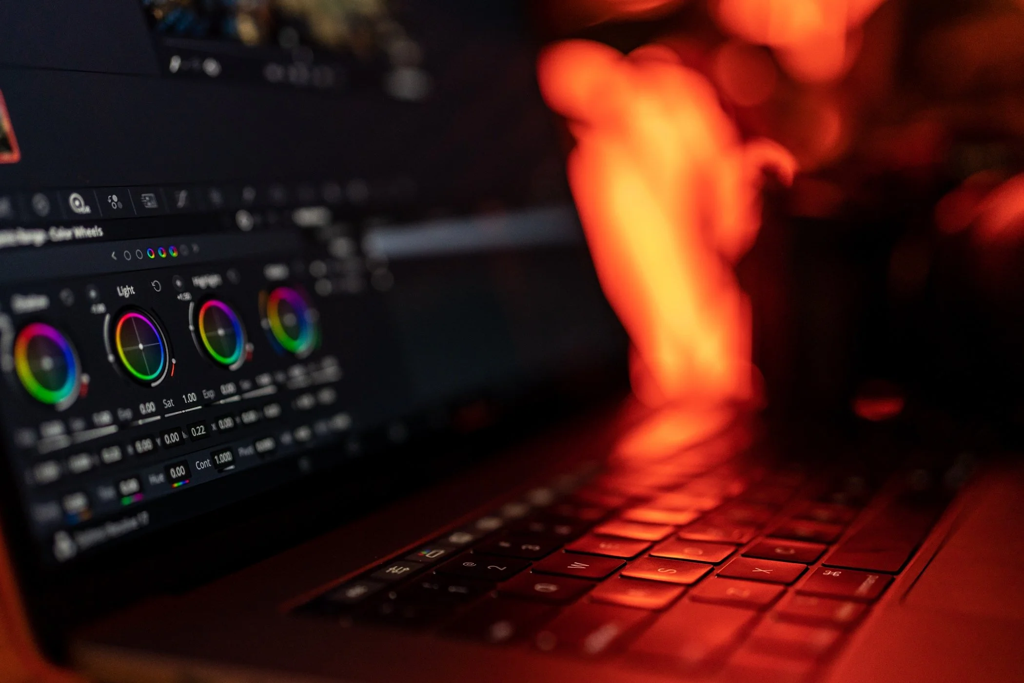 Close-up of a computer keyboard illuminated with orange and red lights, with a laptop screen showing color correction software and fiery orange and red flames in the background.