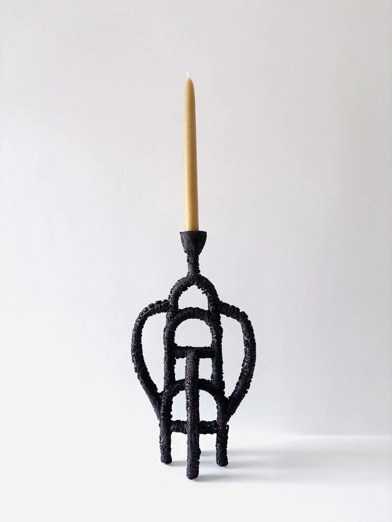 A black, ornate candle holder with a single yellow beeswax candle on top, set against a plain white background.