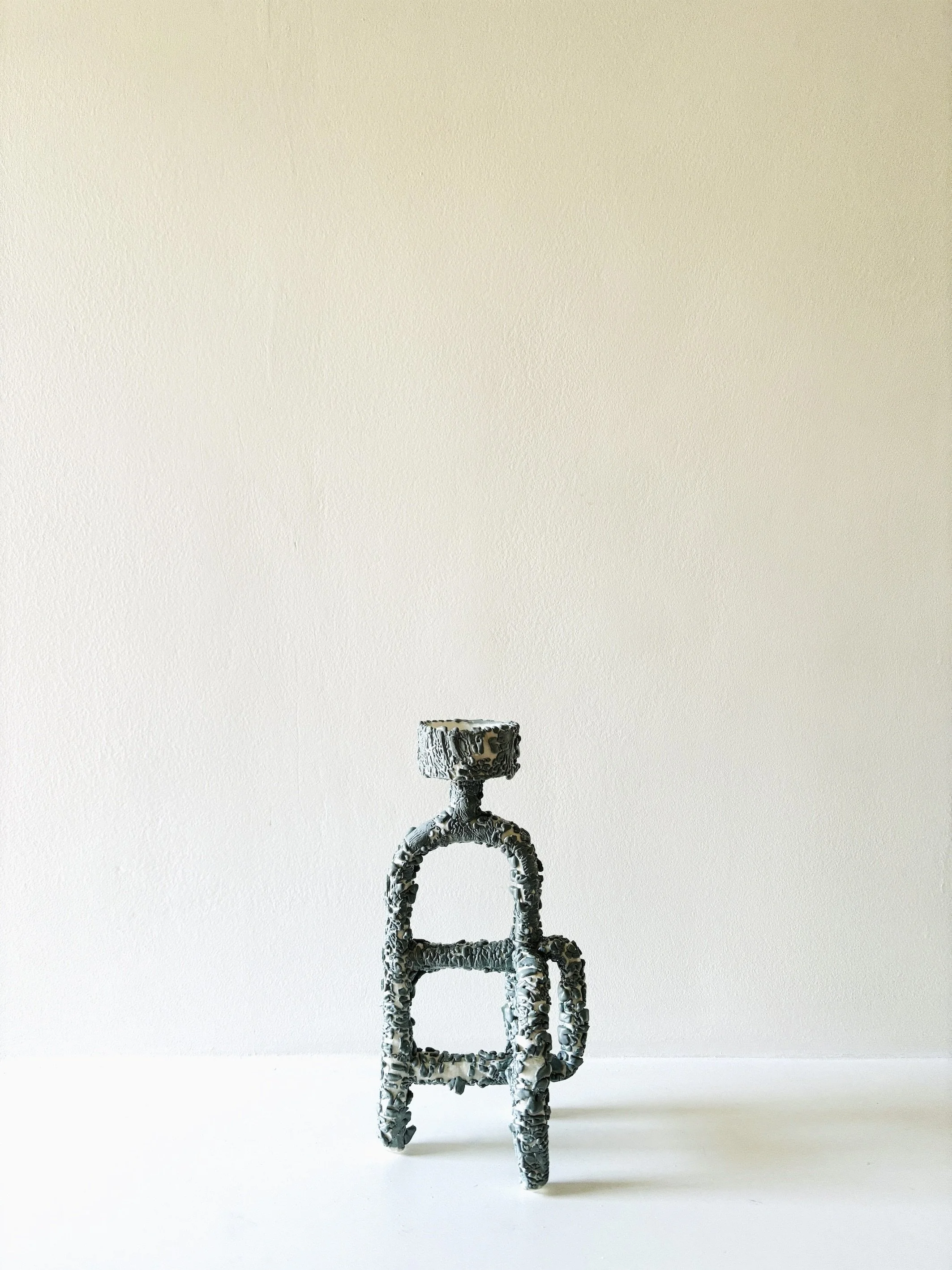 A metallic sculpture resembling an industrial chair, with a textured and intricate design, placed against a plain white wall and on a white surface.