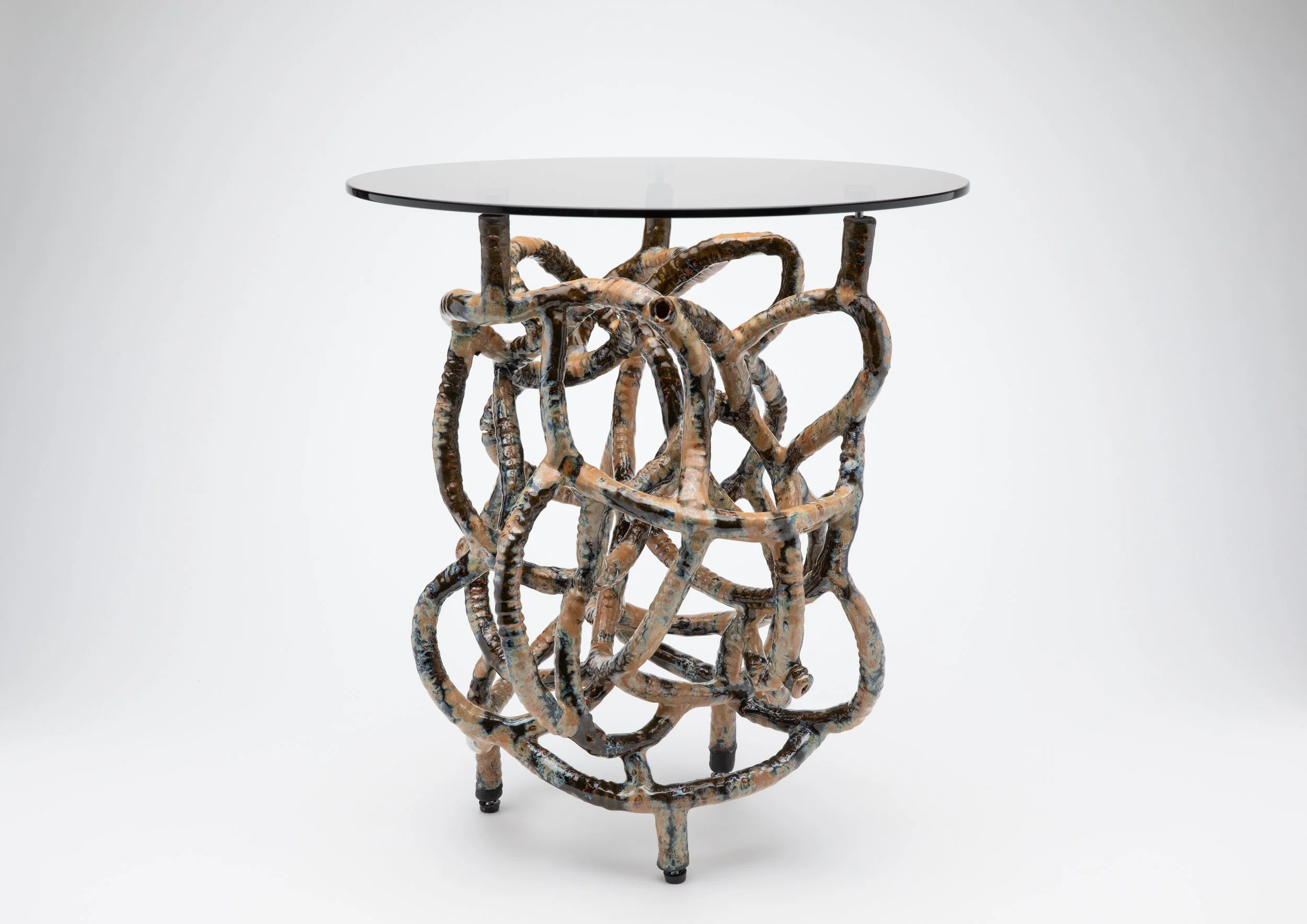 A modern side table with a round glass top and an intricate base made of intertwined, textured, glazed ceramic tubes, standing on three small legs.